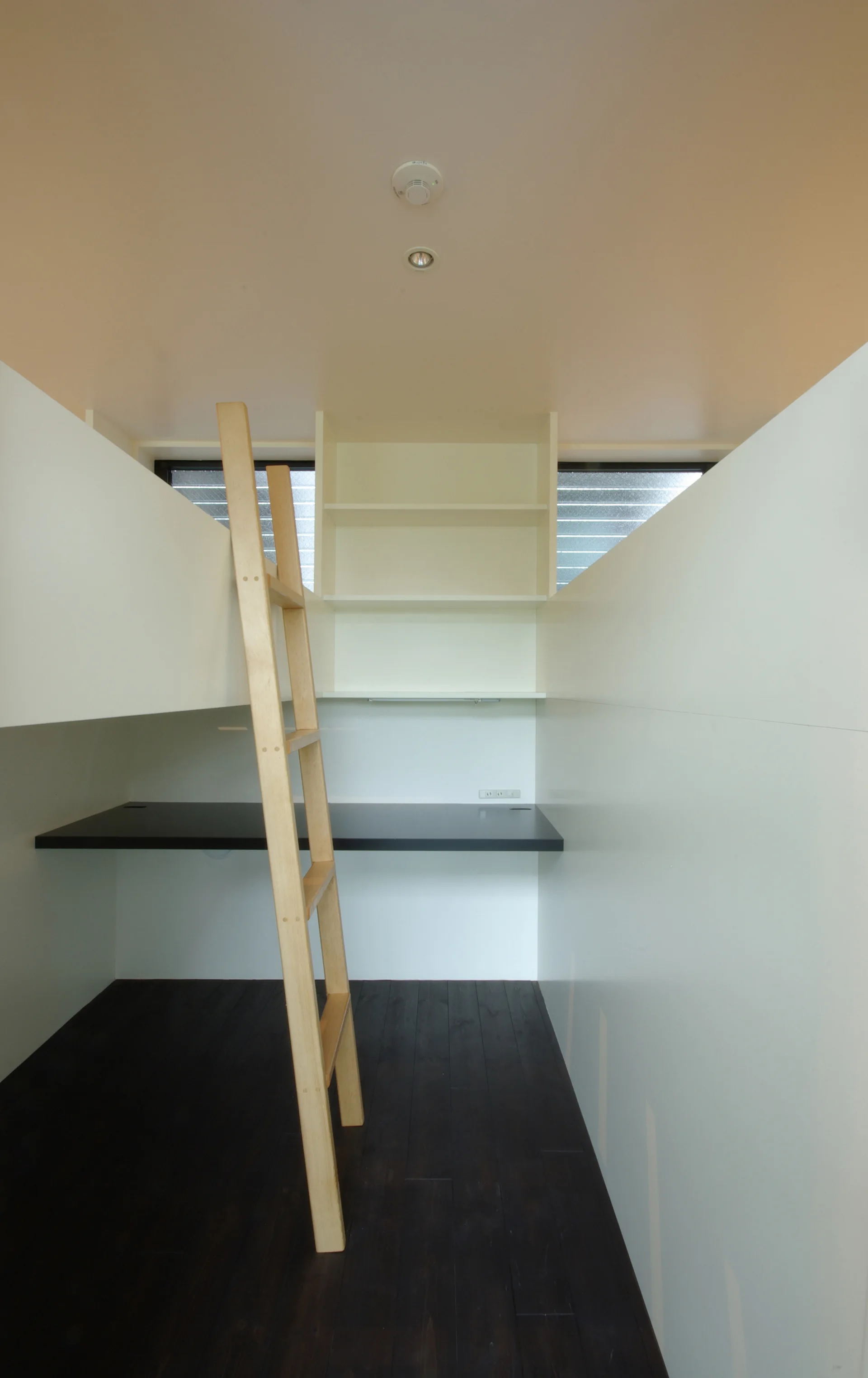 A minimalist compact white loft features a built-in dark desk, white shelves, dark flooring, and a light wood ladder.
