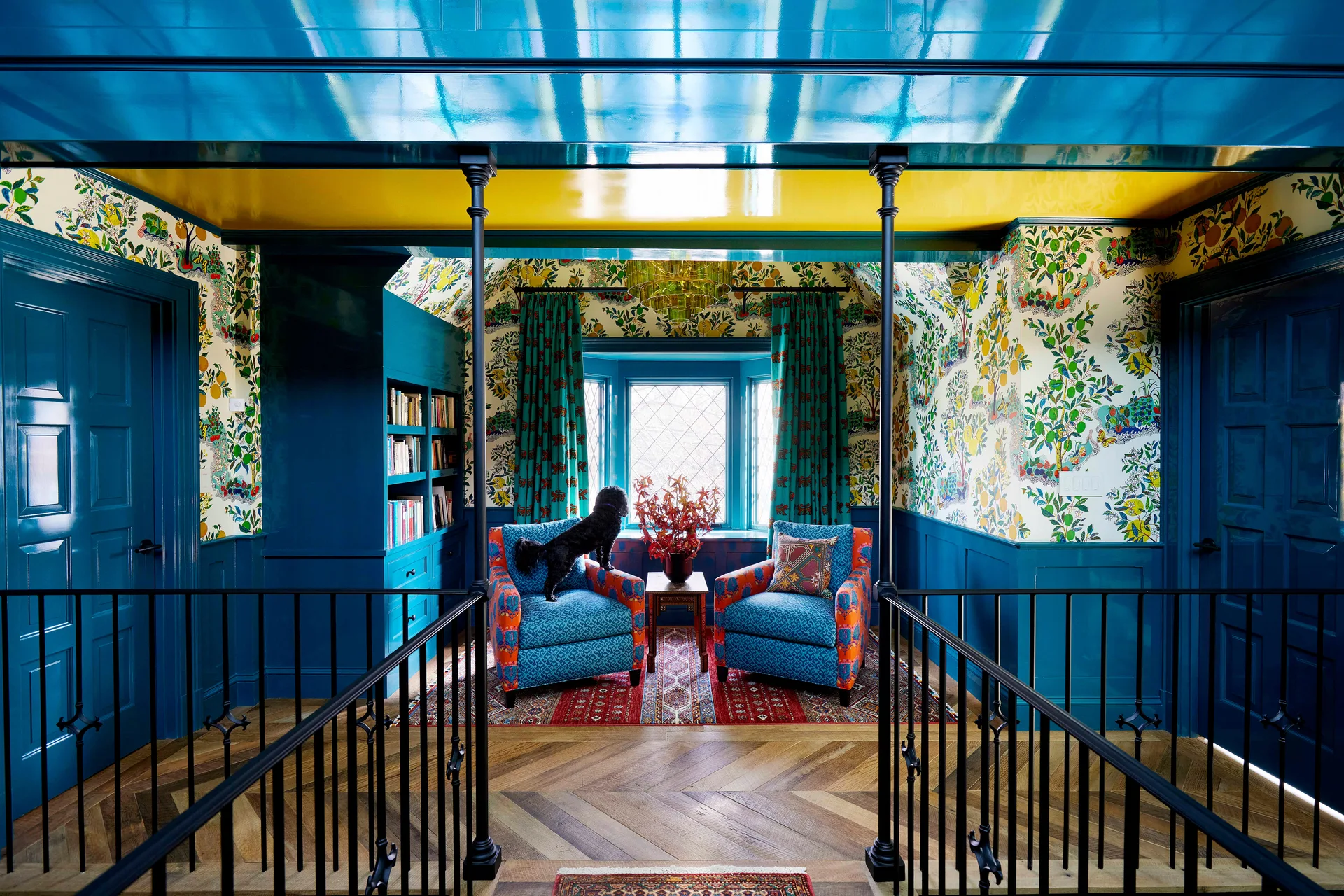 A vibrant landing features a high-gloss blue and yellow ceiling, botanical wallpaper, blue wainscoting, and two patterned blue armchairs on a patterned rug.