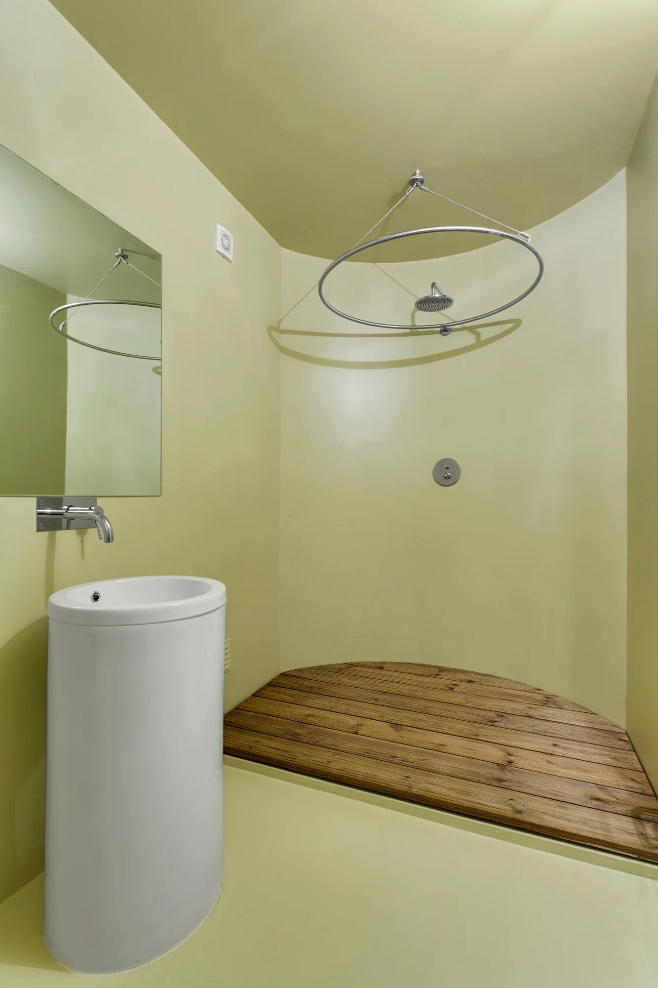 A minimalist bathroom features pale sage green walls, a white pedestal sink, and a curved shower area with a slatted wood floor.