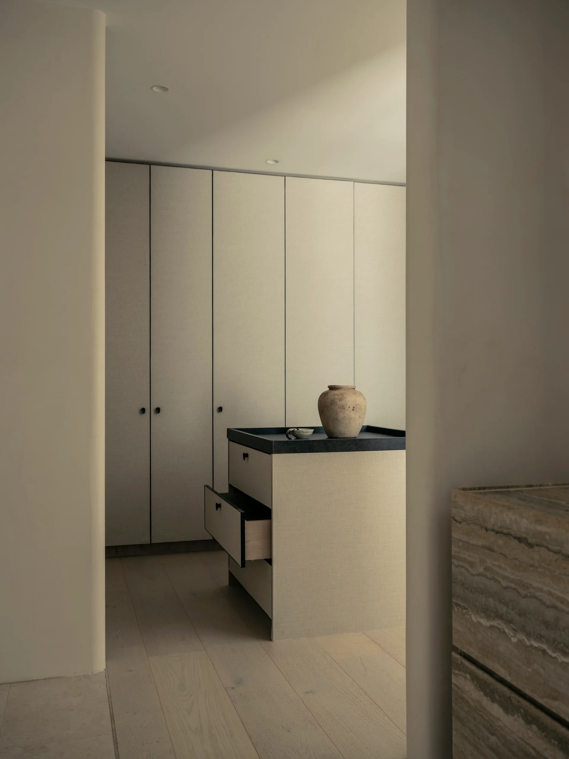 A minimalist dressing room seen through a curved doorway features linen-textured built-in wardrobes and a central storage island with a black top.