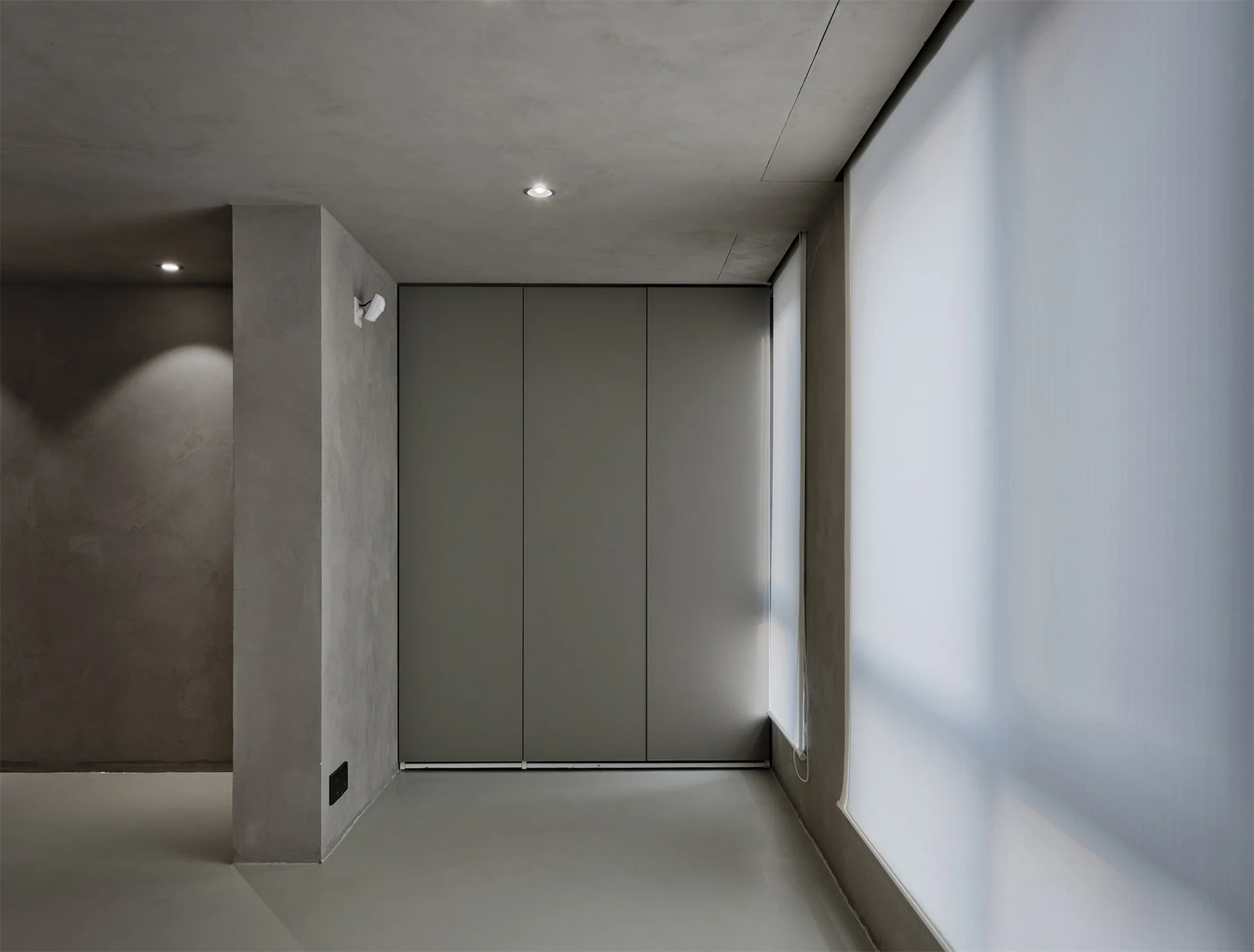 A minimalist gray room features seamless floor-to-ceiling closet doors, textured concrete-like walls, and a large window with a translucent white shade.