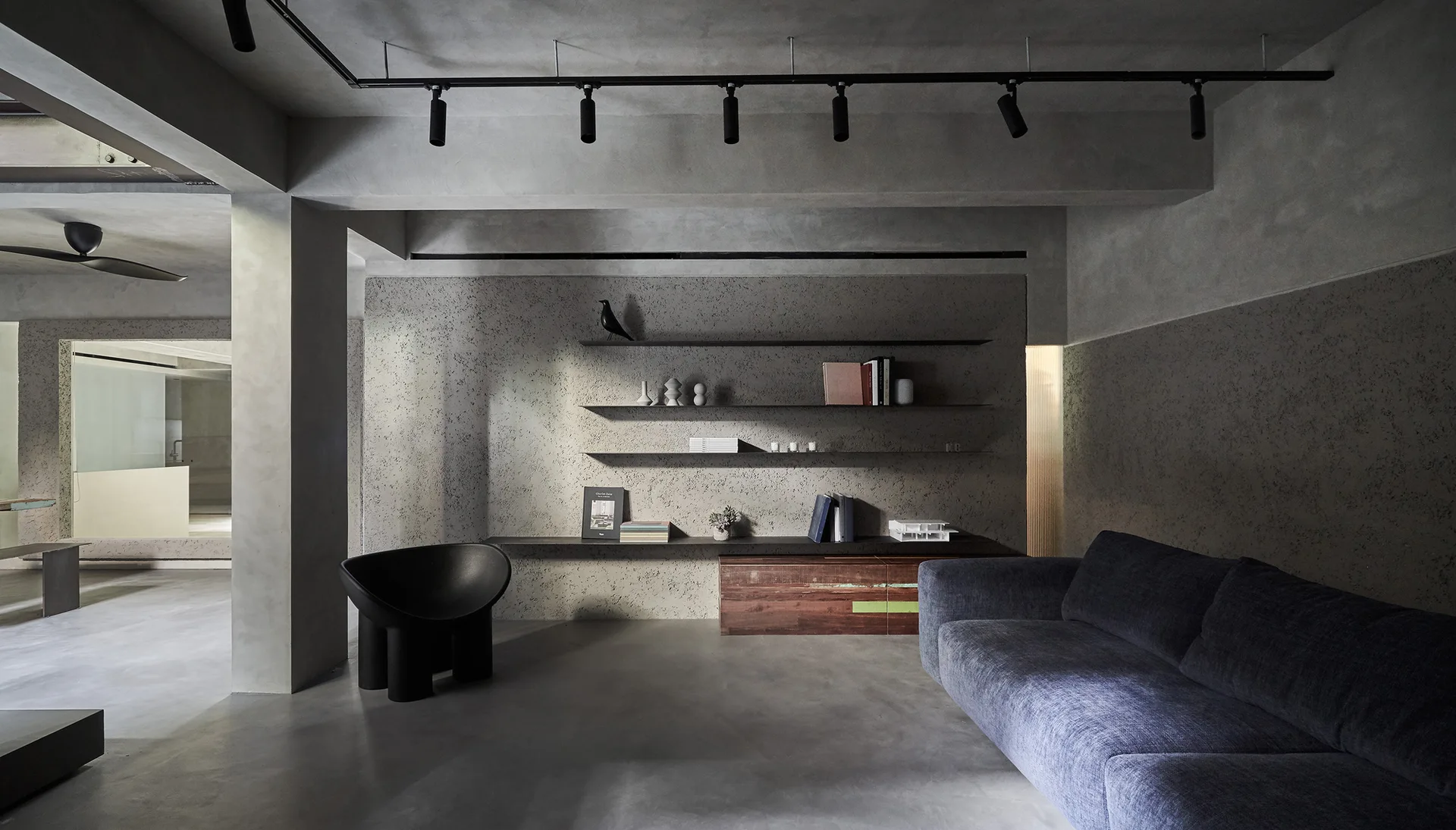 A minimalist grey living room features textured concrete walls, black floating shelves holding books, a sculptural black armchair, and a fabric sofa.
