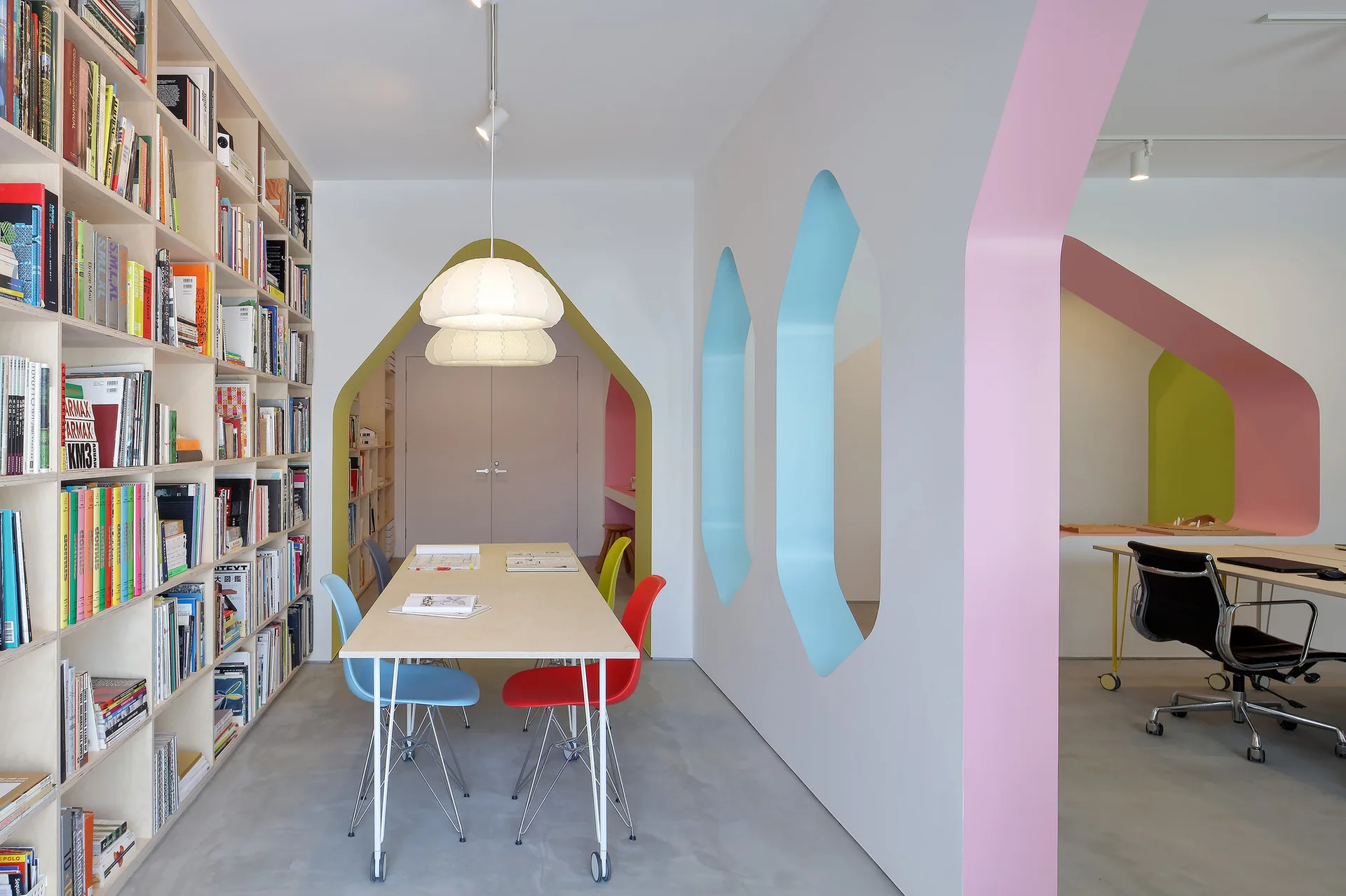 A modern shared workspace featuring a large floor-to-ceiling plywood bookshelf, a wooden desk with colorful chairs, and pastel-painted geometric archways.