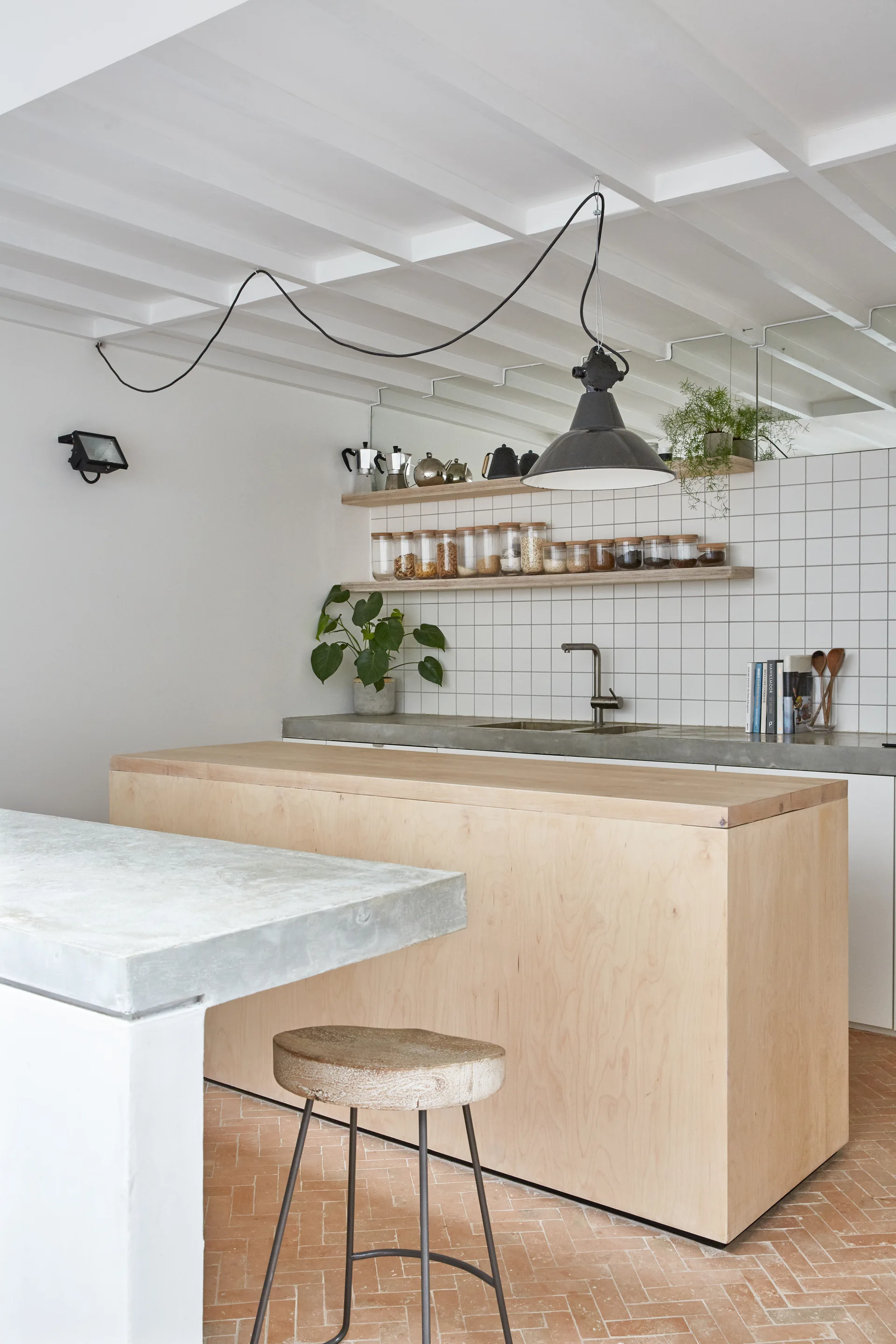A minimalist kitchen features a light plywood island, concrete countertops, a white square tiled backsplash, and a terracotta herringbone brick floor.