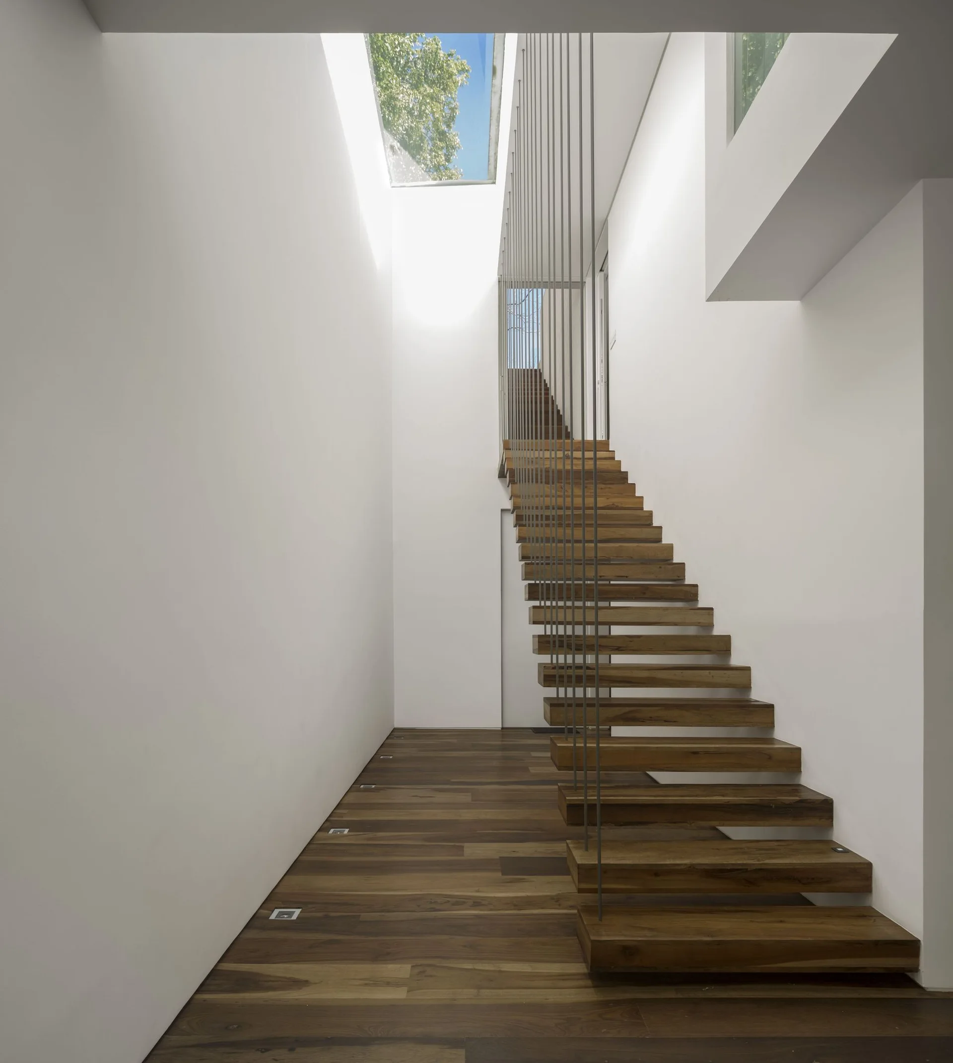 A minimalist floating wooden staircase features vertical cable railings and is illuminated by a skylight in a white hallway.