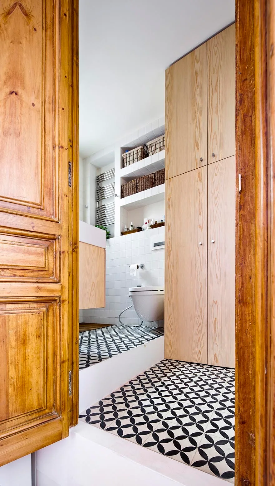 A view through an open wooden doorway into a bathroom with black and white geometric floor tiles, light wood cabinets, and white wall tiles.