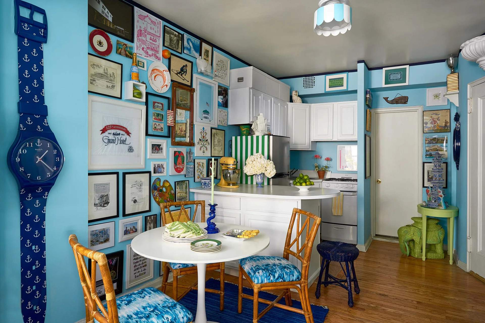 A bright blue dining nook features a white round table, rattan chairs with blue cushions, and a dense gallery wall next to white kitchen cabinets.
