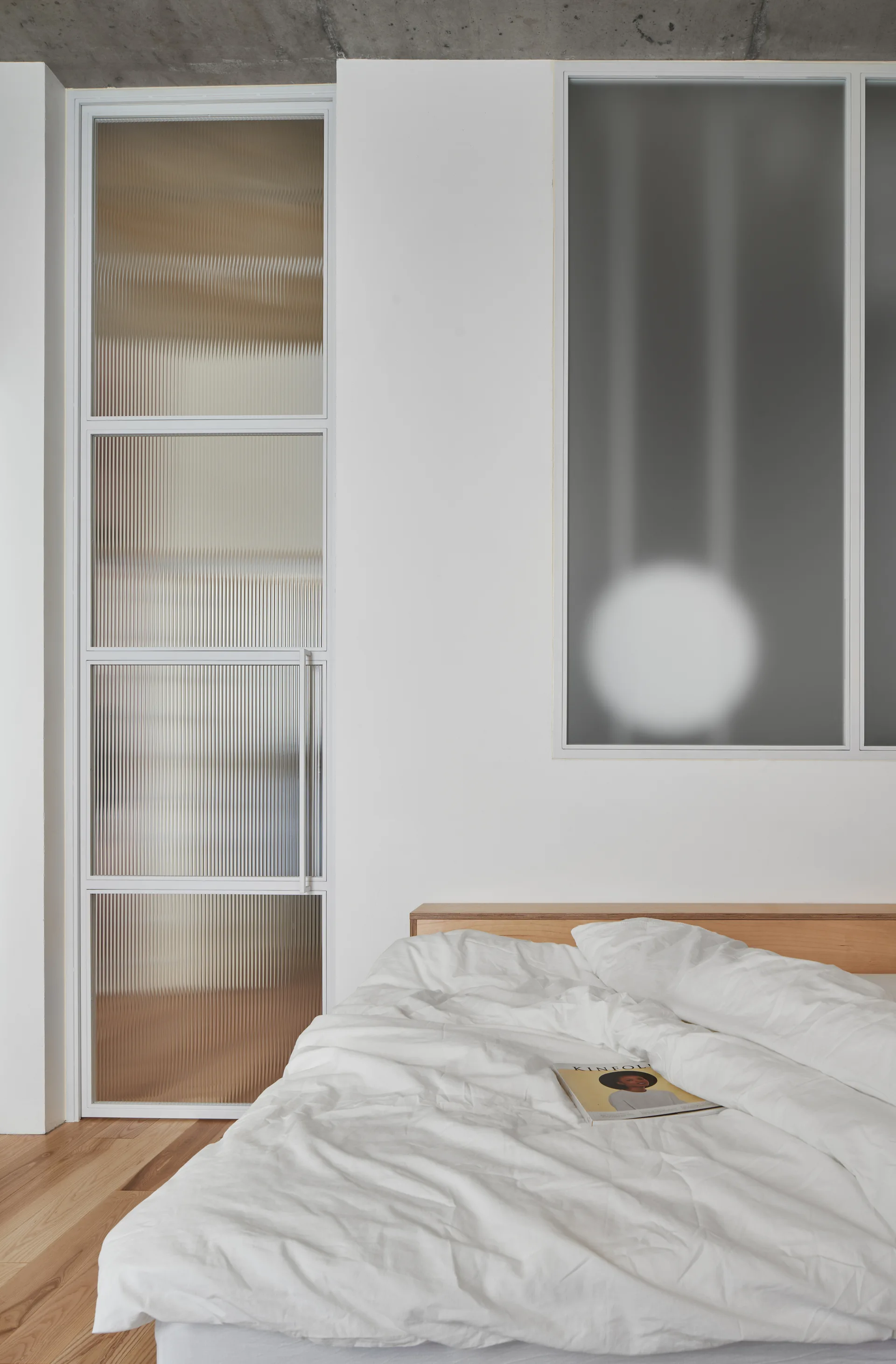 A minimalist bedroom featuring a bed with white bedding and a wood headboard next to a fluted glass partition and a grey concrete ceiling.