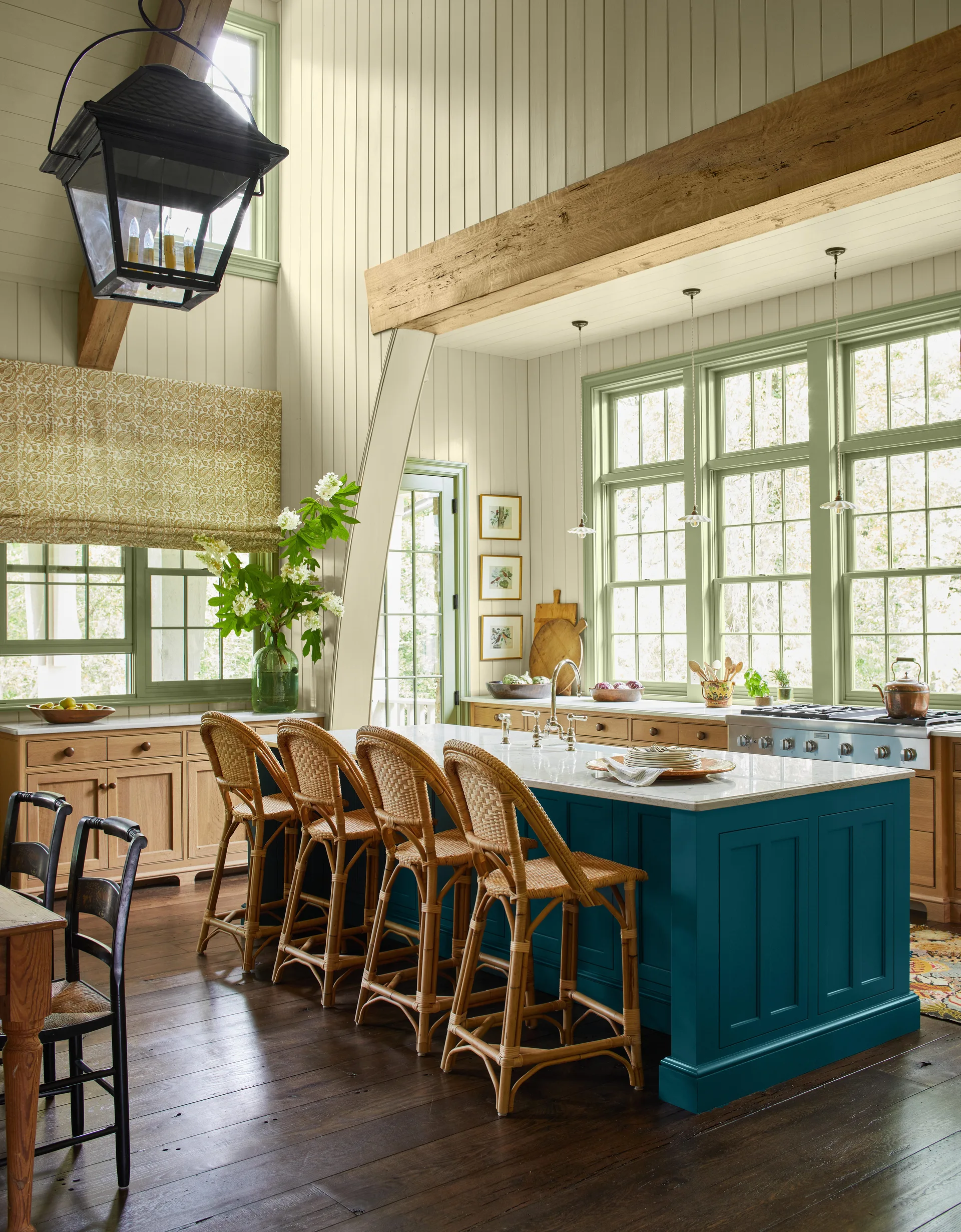 A farmhouse kitchen features a teal island with rattan bar stools, vertical cream wall paneling, and windows trimmed in sage green.