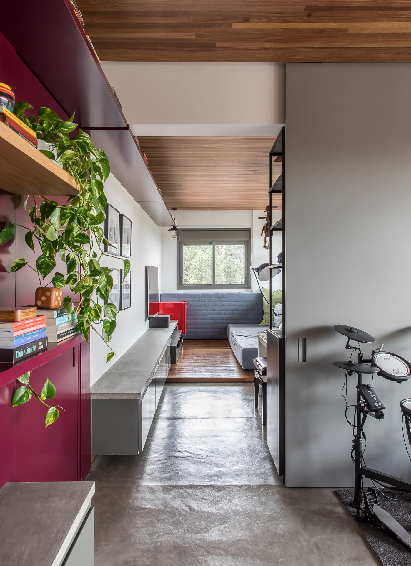 A modern narrow room featuring a magenta built-in shelving unit, a concrete floor, a wood-paneled ceiling, and a gray sliding door.