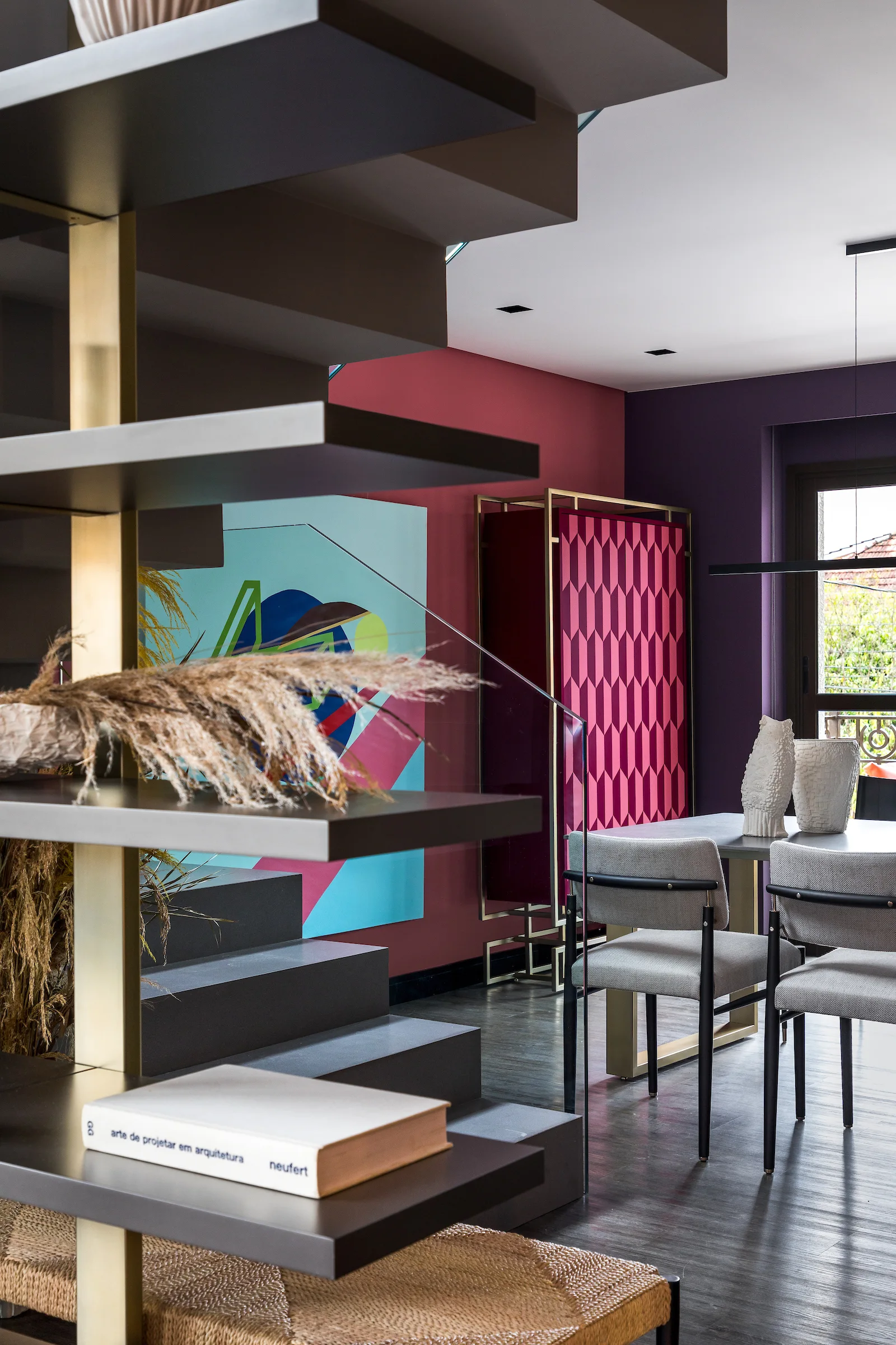 A modern dining space features a dark sculptural staircase with built-in shelves next to a purple and pink patterned cabinet.