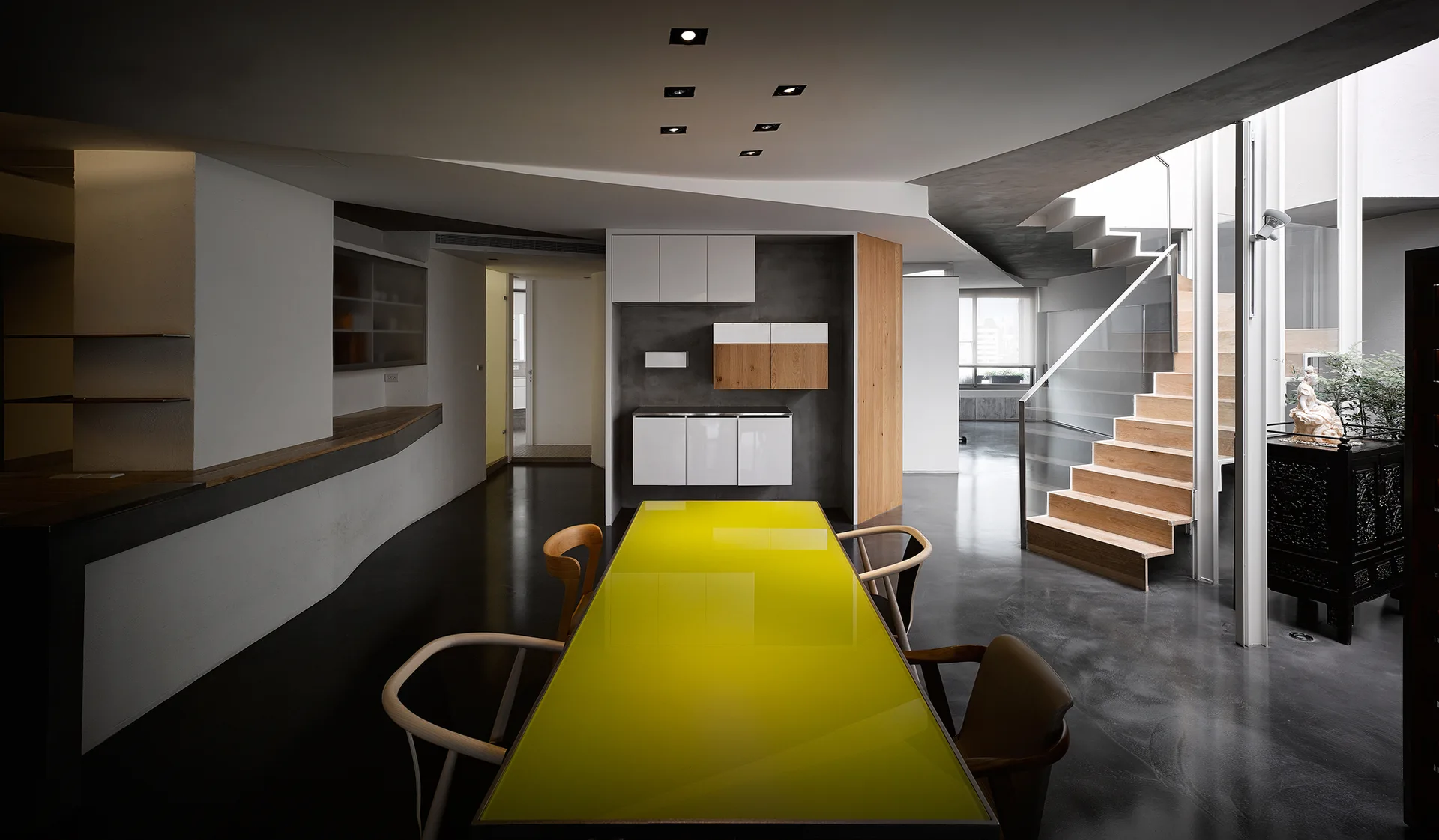A modern dining room features a long table with a bright yellow top, surrounded by wooden chairs, set on a dark concrete floor next to wooden stairs.