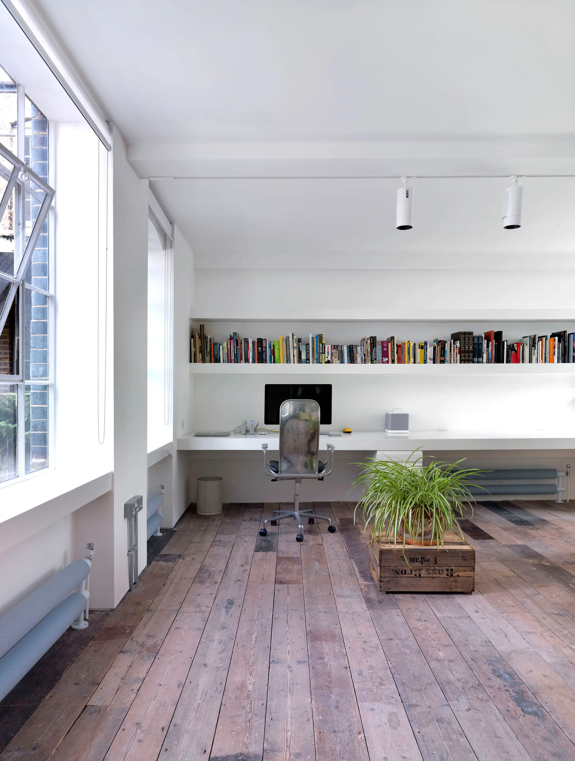 A minimalist home office featuring a long wall-integrated white desk and bookshelf set over rustic, weathered wooden floorboards.