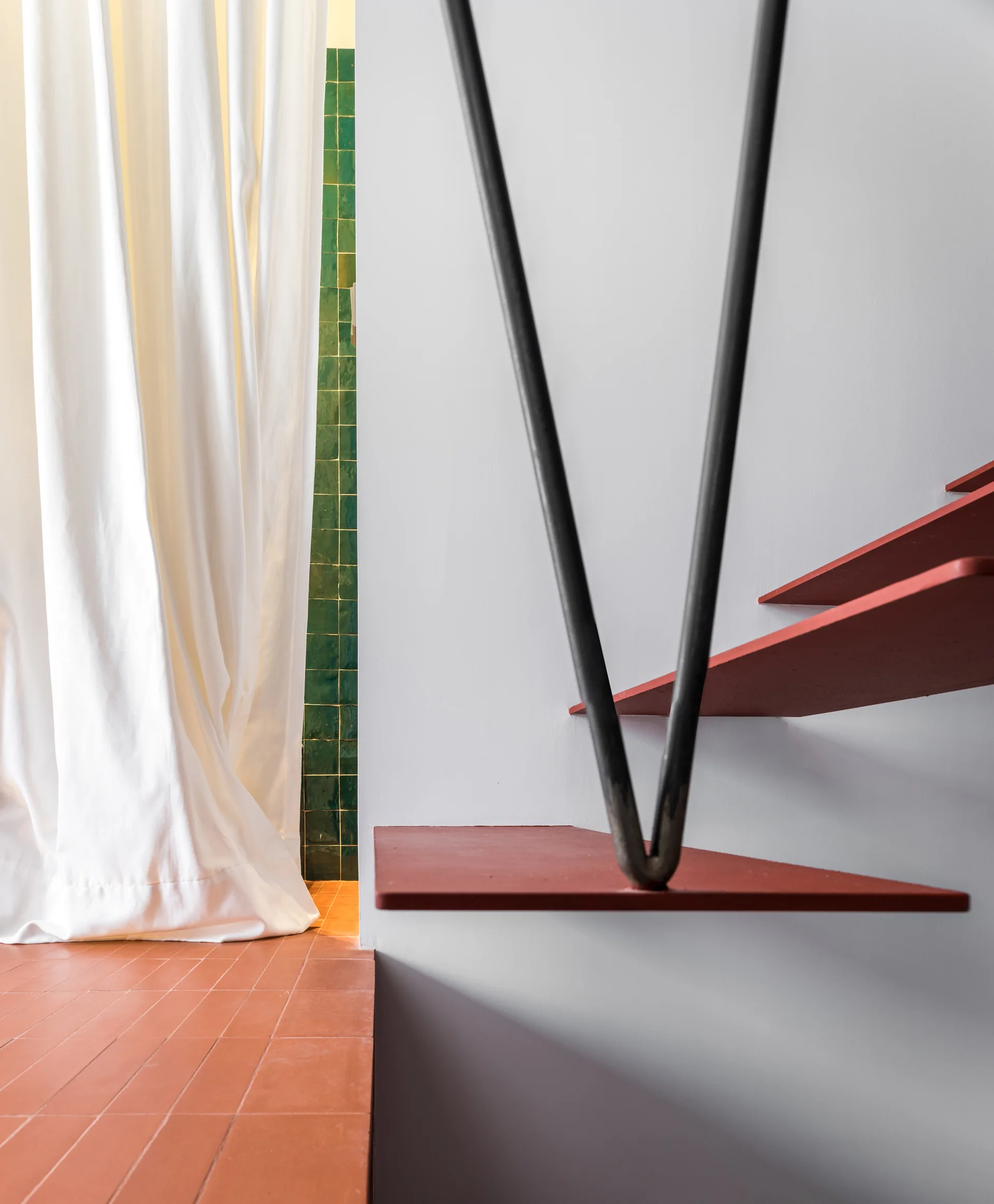 Minimalist red floating stair treads are suspended beside a white wall, adjacent to a terracotta floor, a white curtain, and green wall tiles.