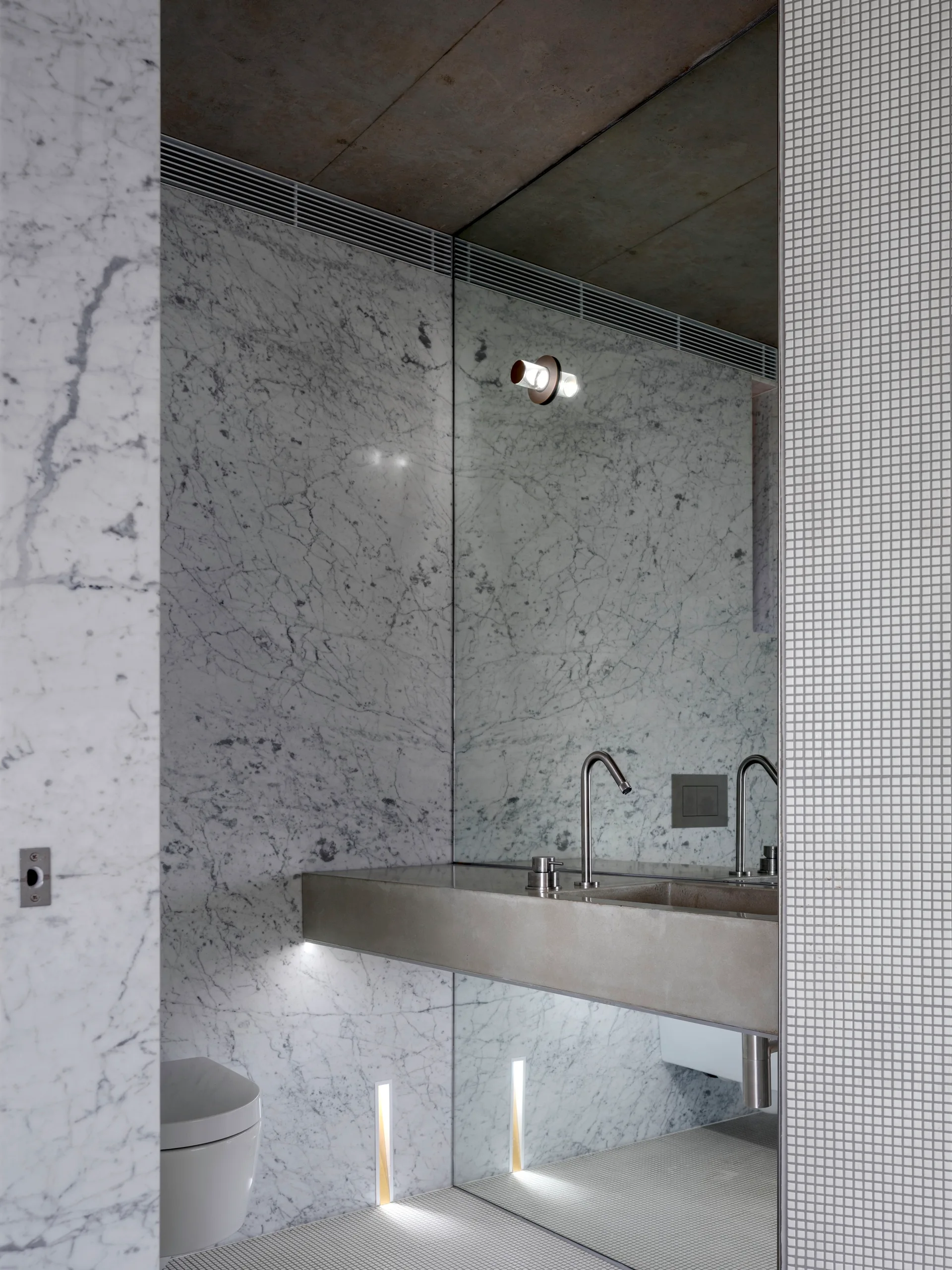 A minimalist bathroom features a wall-mounted metal vanity, white veined marble walls, a full-height mirror, and small mosaic grid tiles.