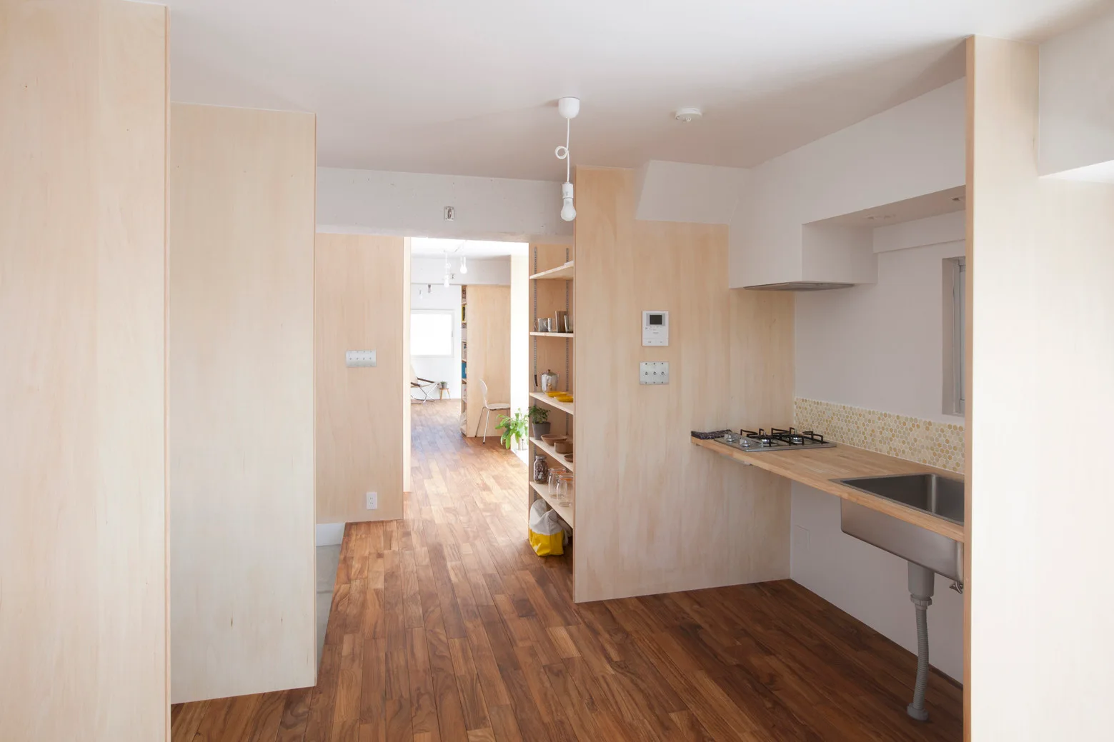 A minimalist kitchen features light plywood wall paneling, a wooden countertop with a stainless steel sink, and dark wood plank flooring.