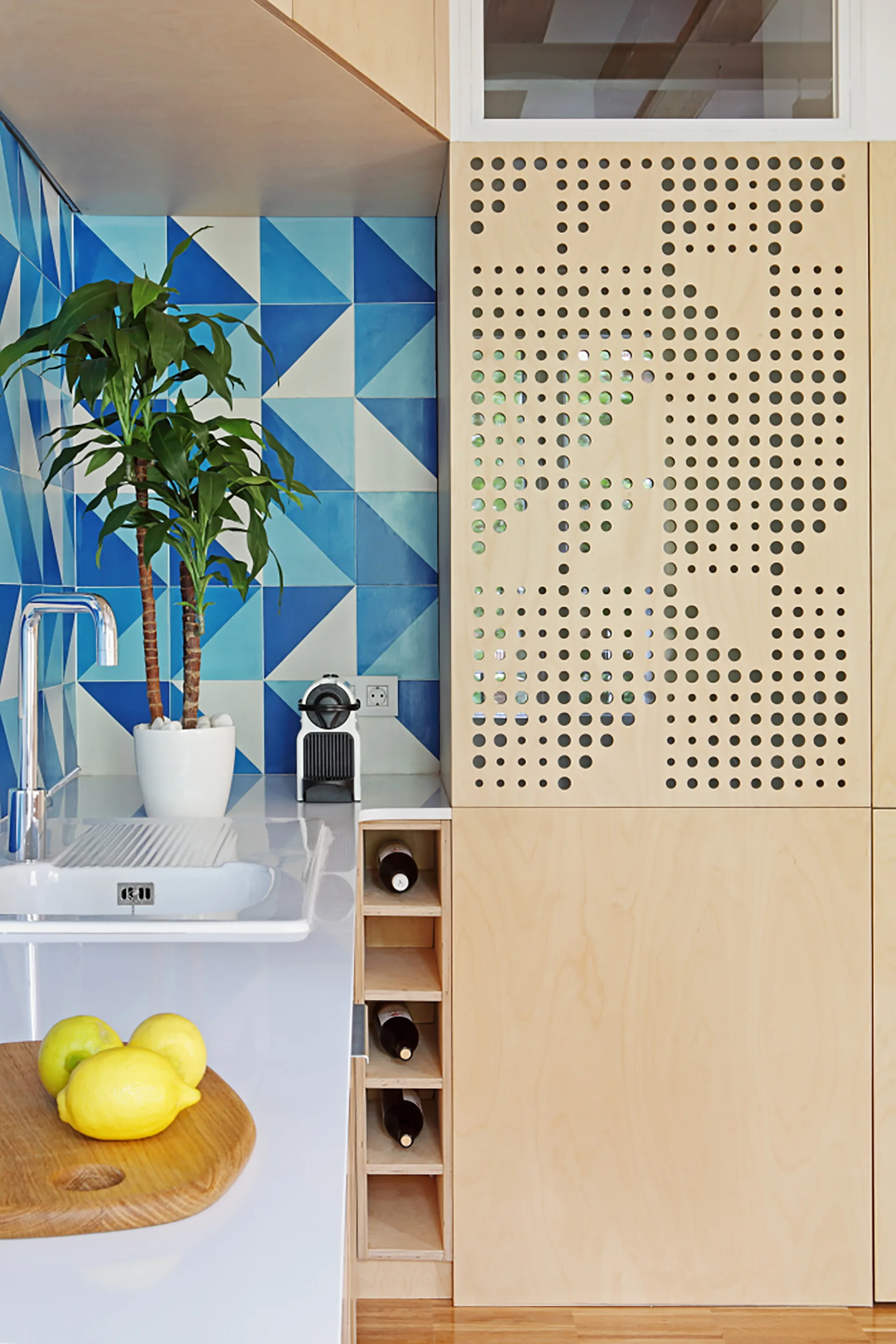 A modern kitchen corner features a light wood cabinet with perforated patterns, a blue triangular tile backsplash, and an integrated white sink.