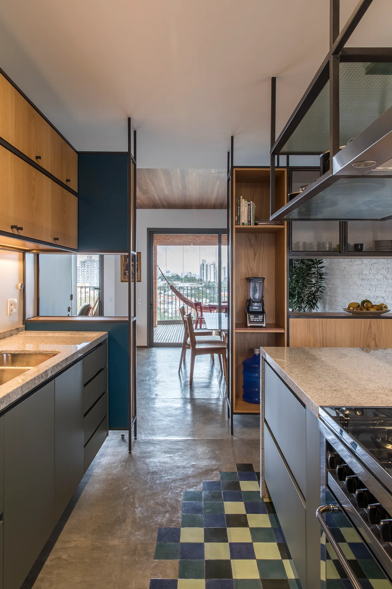 A galley kitchen features dark grey cabinets, wood accents, a black metal shelving unit, and a checkered tile floor inlay leading toward a balcony.