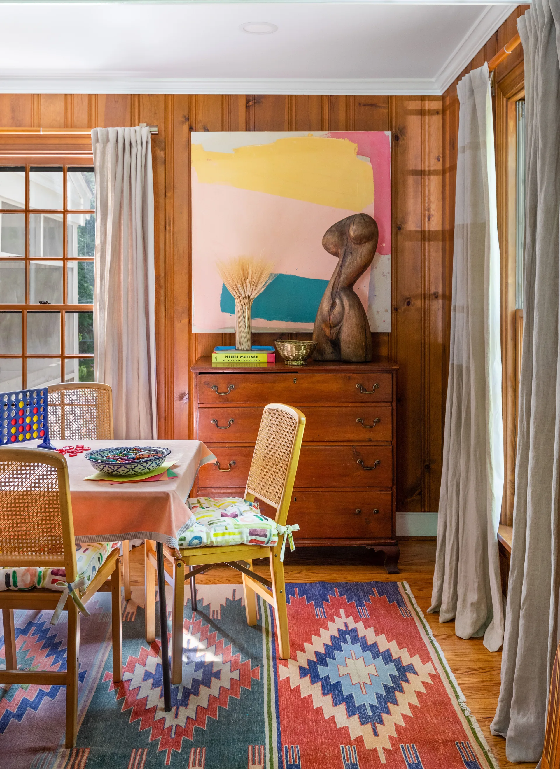 A dining room features vertical wood paneling, cane-back chairs, a geometric patterned rug, and a large colorful painting above a wooden chest.