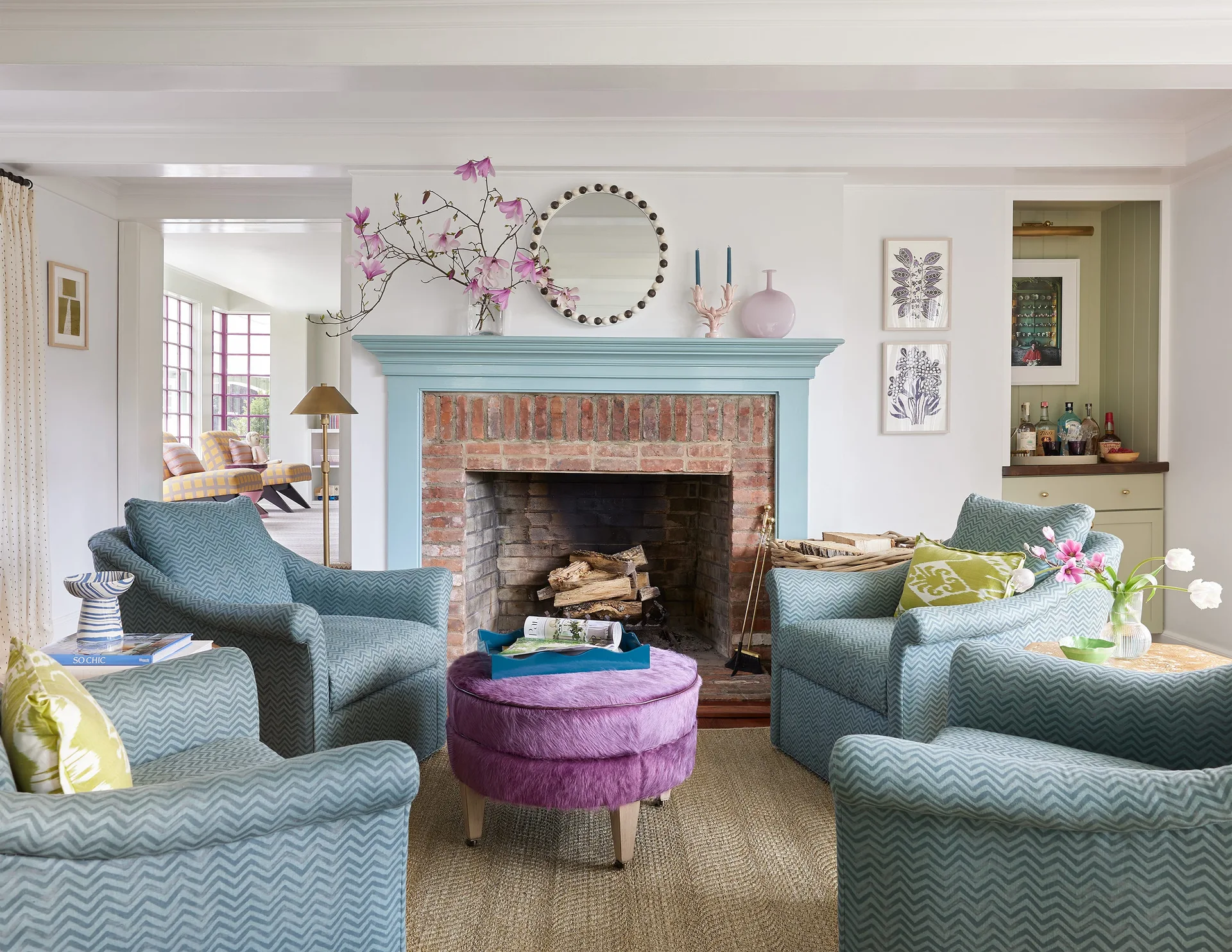 Four blue chevron armchairs are arranged around a purple ottoman in front of a brick fireplace with a painted blue mantel.