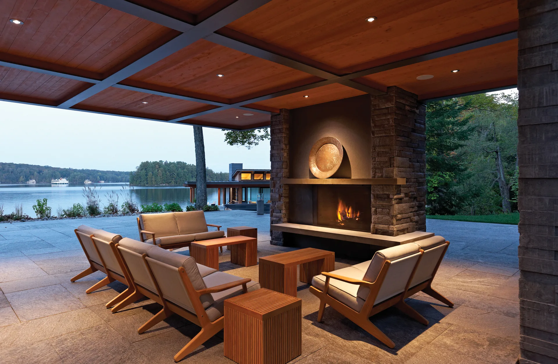 A covered outdoor patio features a stone fireplace, wood-framed sofas, and slatted side tables overlooking a scenic lake.