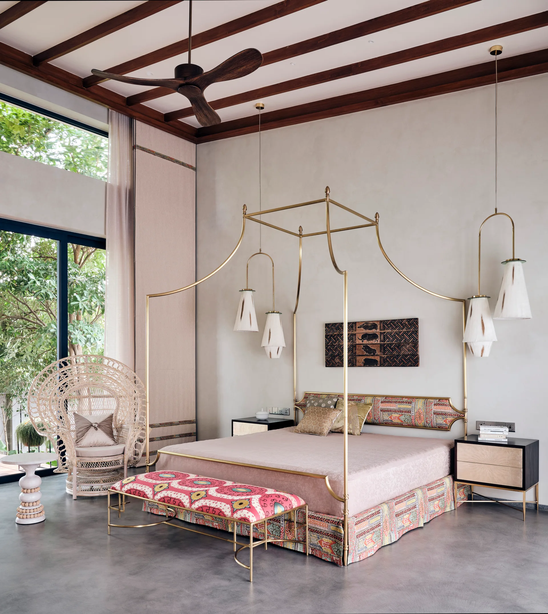 A light gray bedroom features a brass canopy bed, a patterned bench, a woven rattan peacock chair, and exposed dark wood ceiling beams.