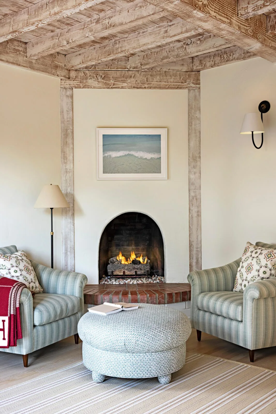 Two striped armchairs and a patterned round ottoman sit before an arched fireplace under a whitewashed wood beamed ceiling.
