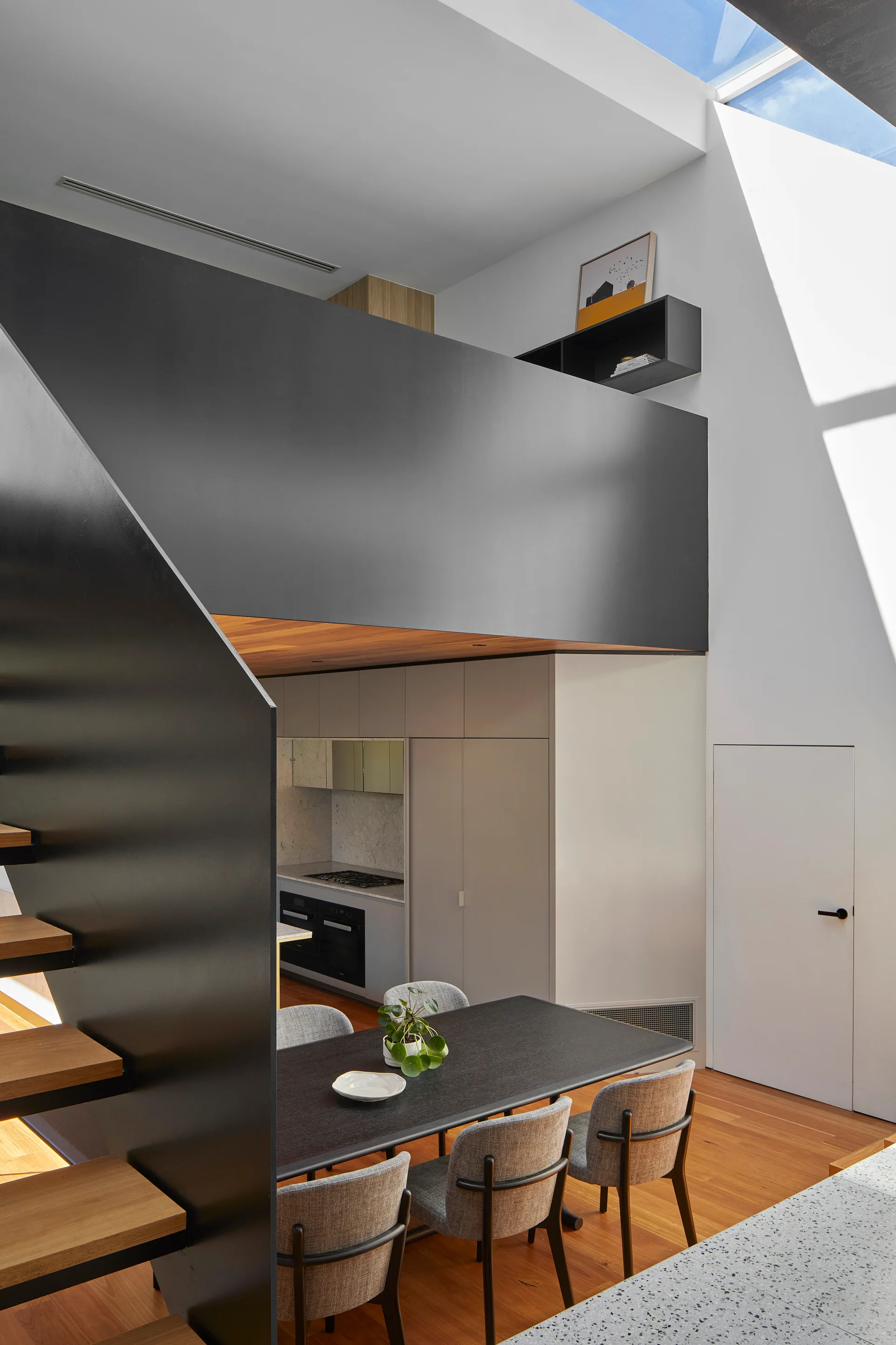 A modern dining room features a black table, grey chairs, and a dark staircase next to a white wall illuminated by an overhead skylight.