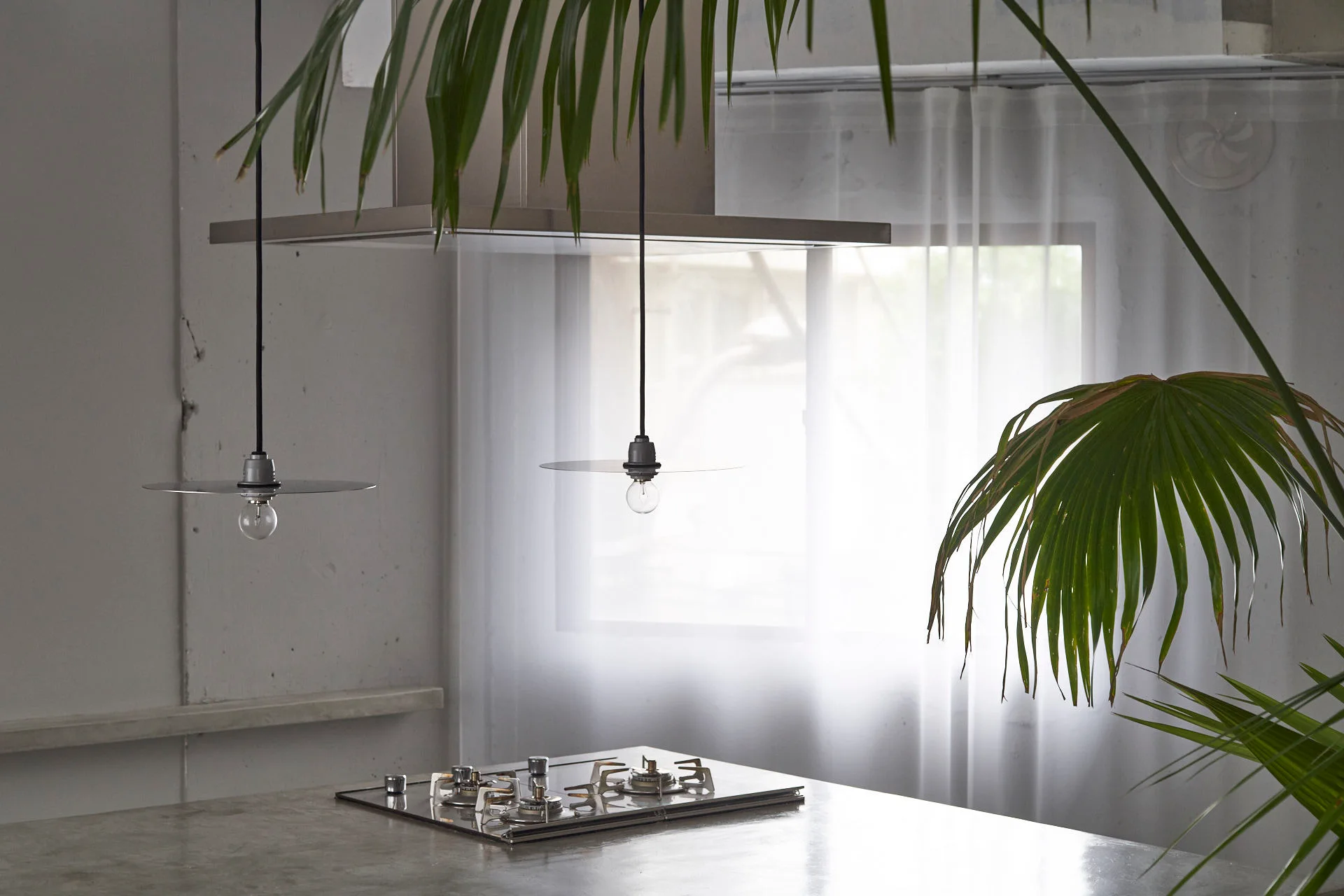 Two minimalist disc pendant lights hang over a concrete kitchen counter with a built-in cooktop, set against a concrete wall and window.