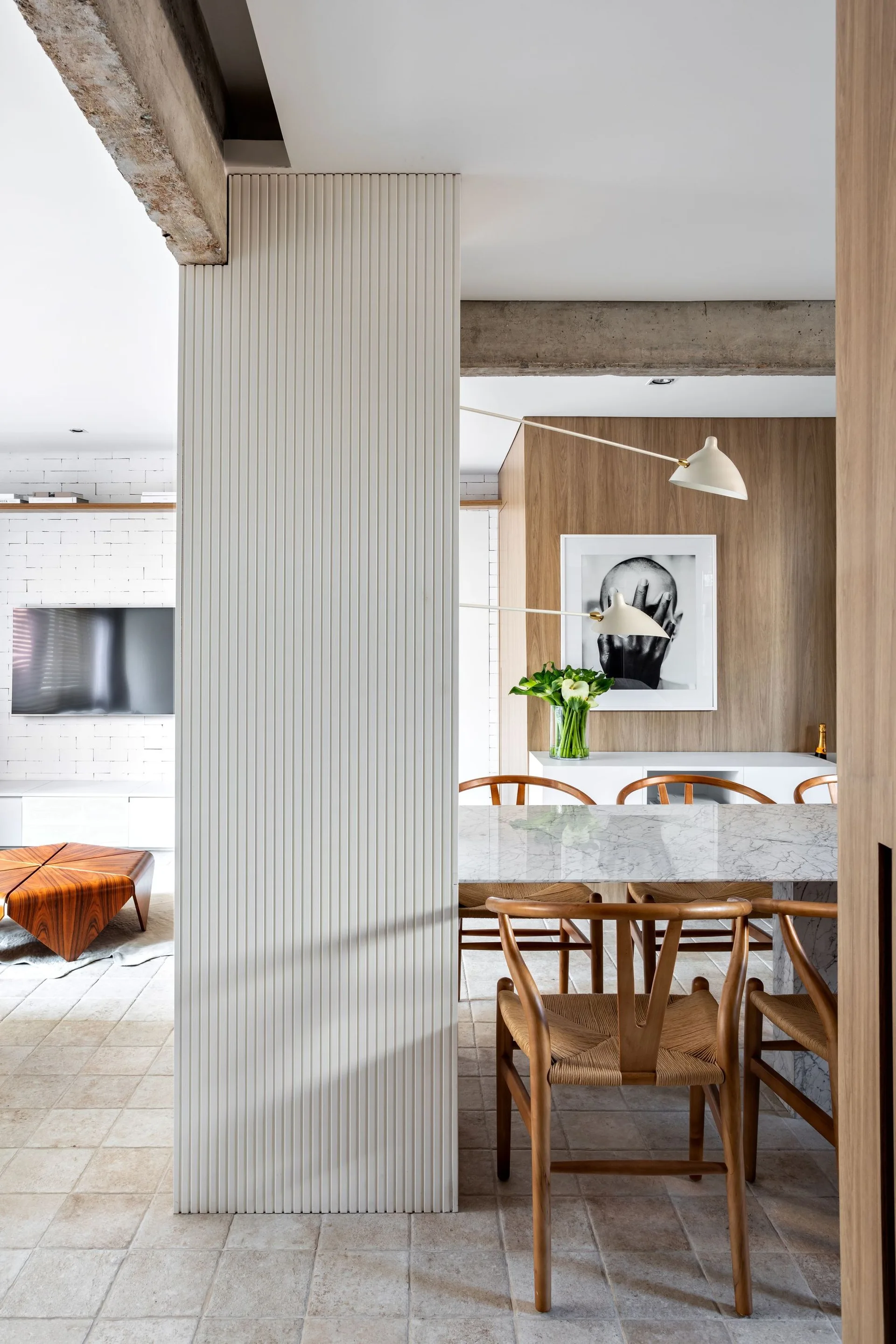 A dining area features a marble-topped table and wood chairs next to a white fluted pillar, with raw concrete beams overhead and a wood-paneled wall.