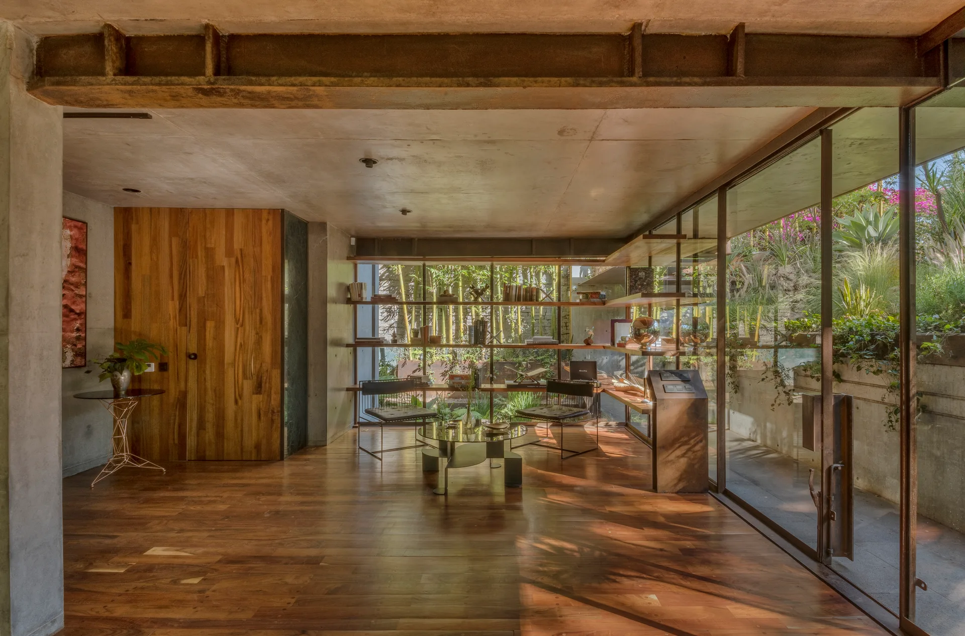 A modern living room features wood floors, exposed concrete ceilings with a steel beam, and floor-to-ceiling windows overlooking a bamboo garden.