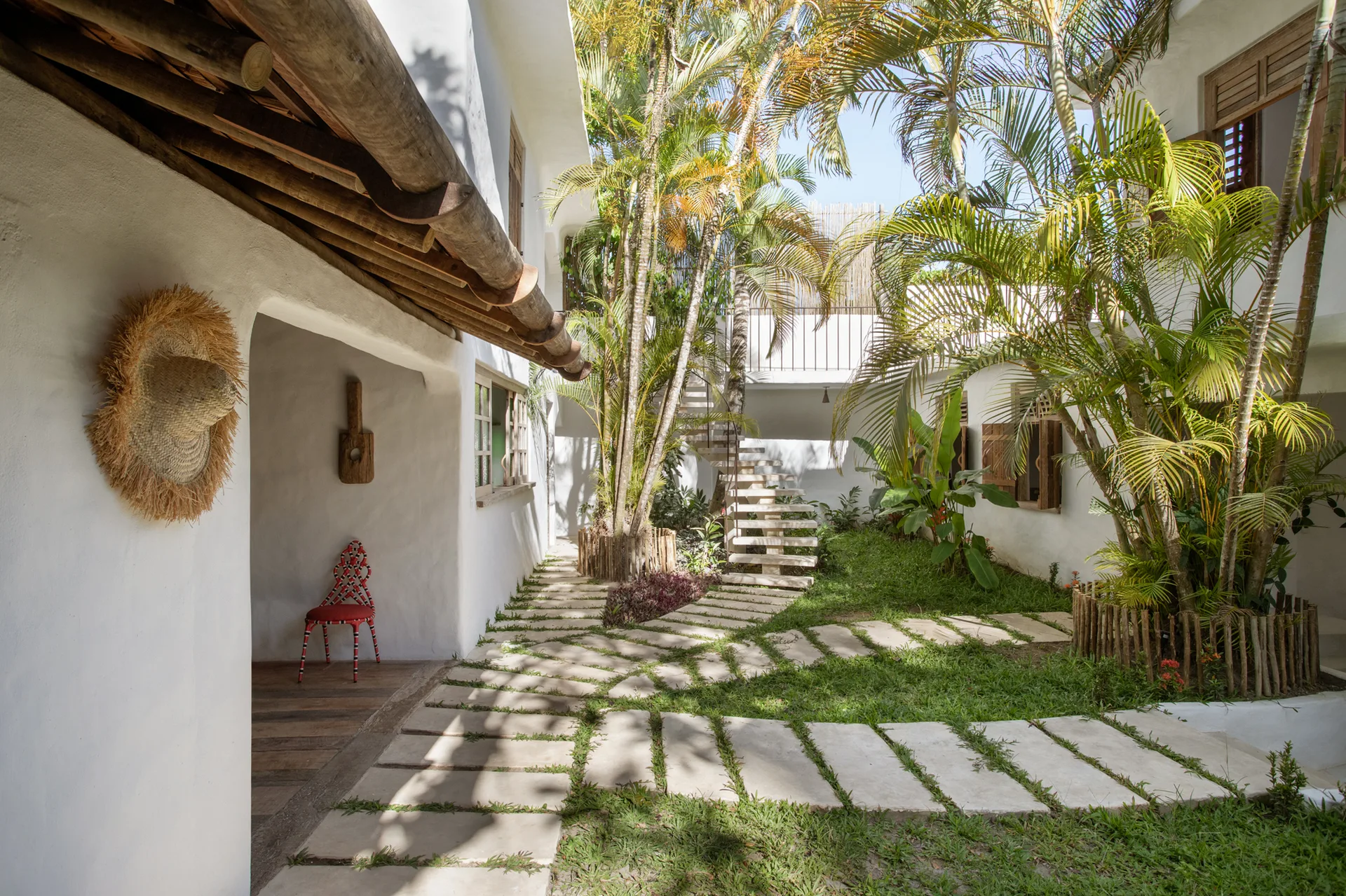 A tropical outdoor courtyard features white stucco walls, a curved stone stepping-path through grass, palm trees, and a floating stone staircase.