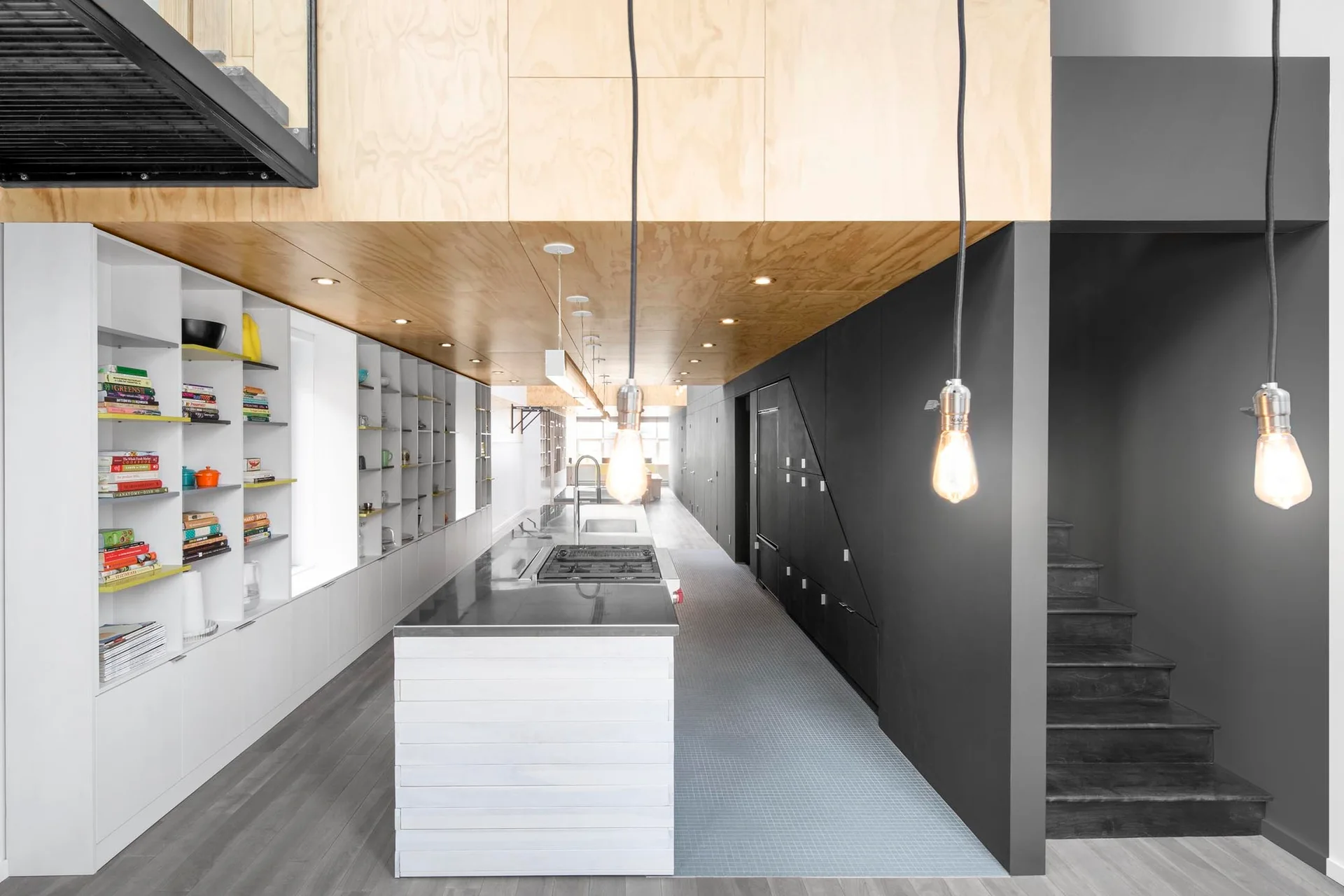 A modern linear kitchen features a central island, a natural wood plywood ceiling panel, white open shelving, and a matte black storage wall.