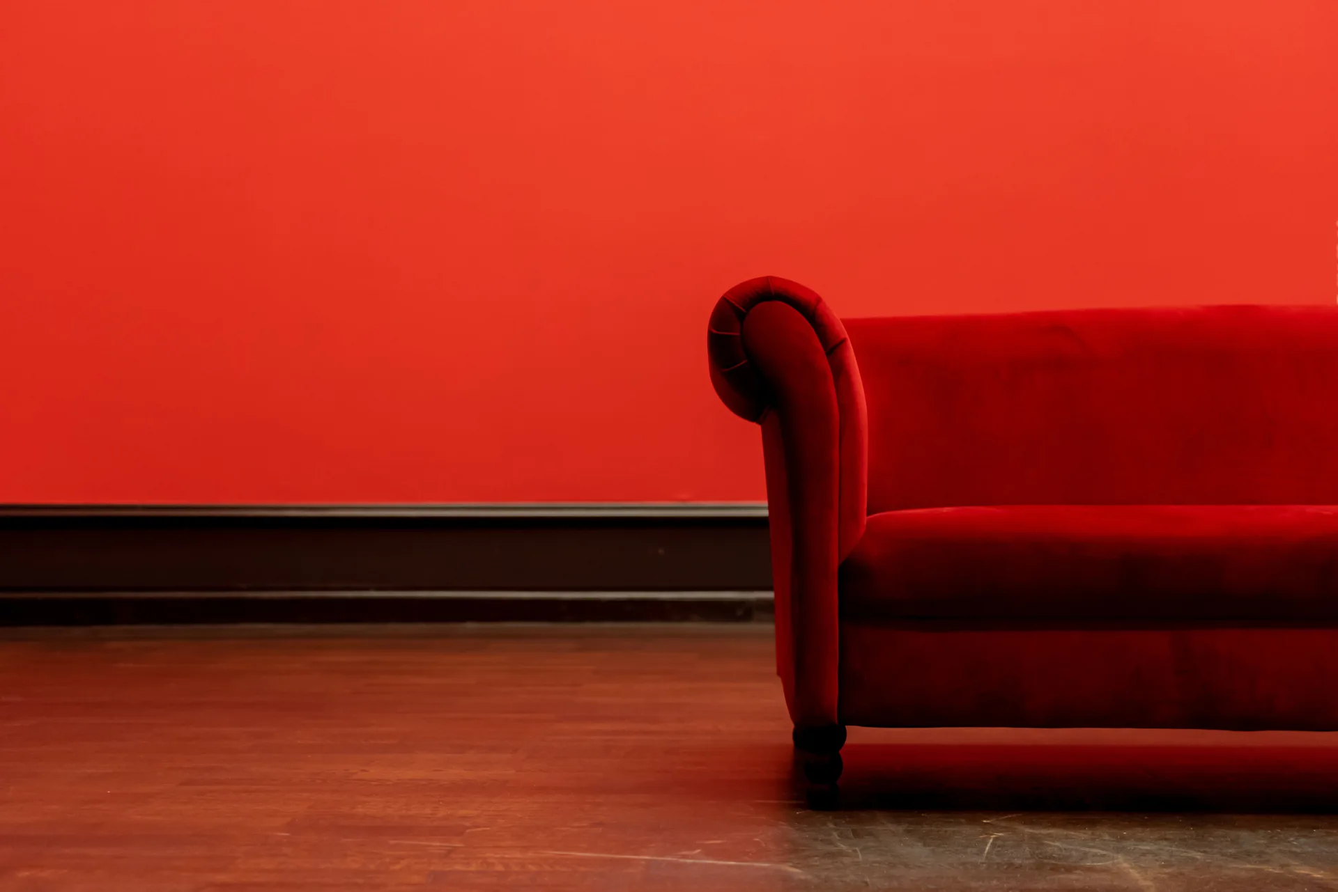 A red velvet sofa with a rolled arm sits on a wood floor against a matching red wall with dark baseboard trim.