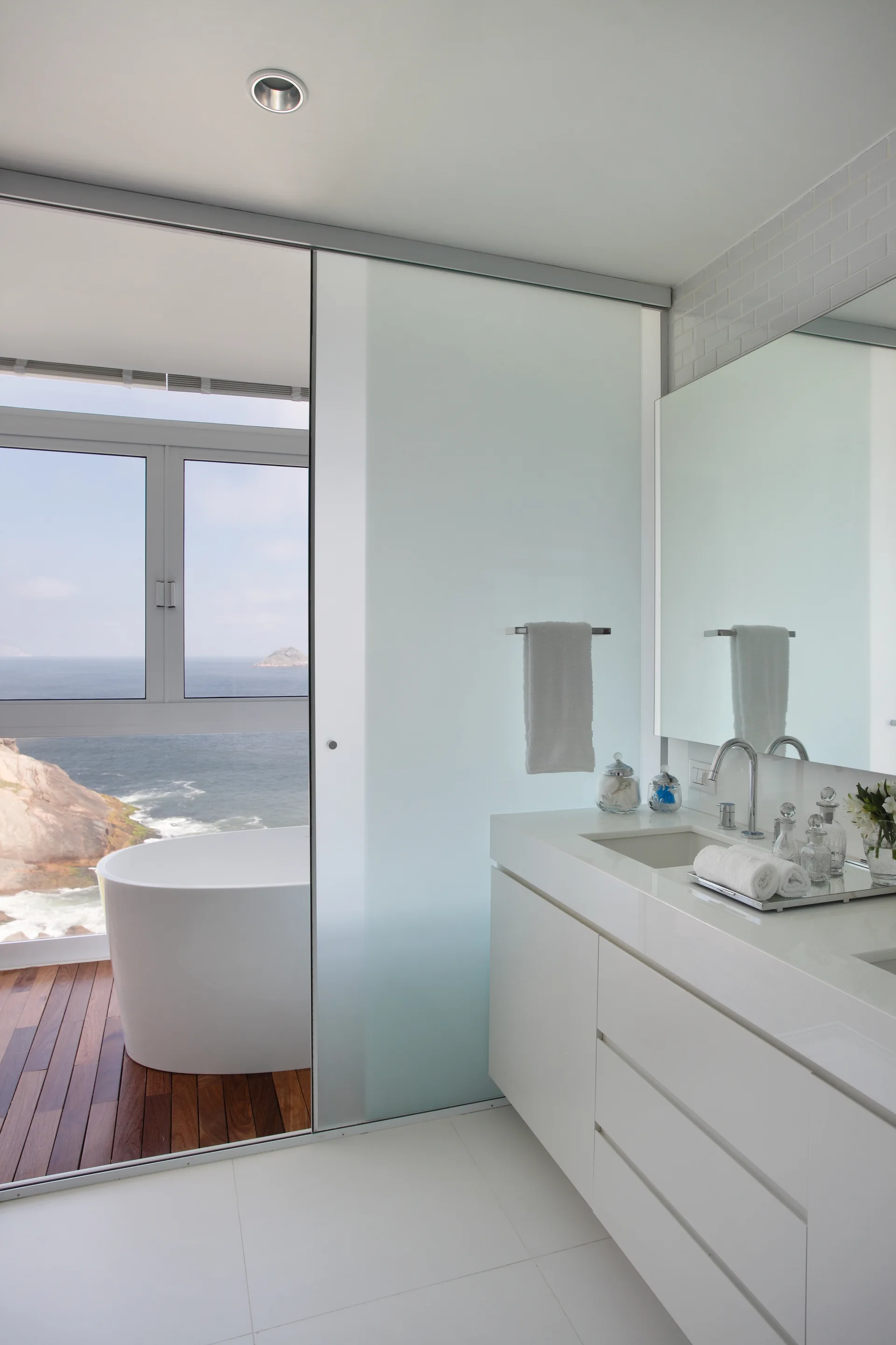 A modern white bathroom features a glossy vanity, a frosted glass sliding door, and a freestanding bathtub set on wood flooring with an ocean view.