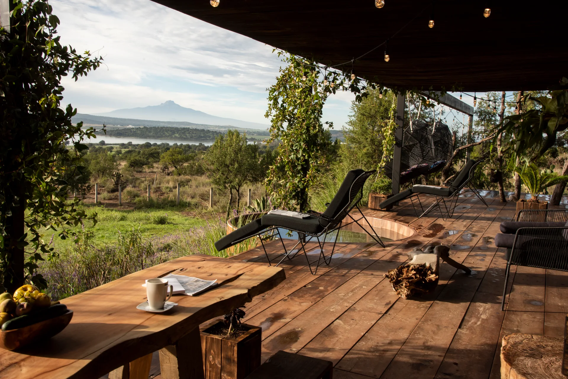 A covered wood deck features black metal lounge chairs, a live-edge wooden table, and a circular hot tub overlooking a scenic mountain landscape.