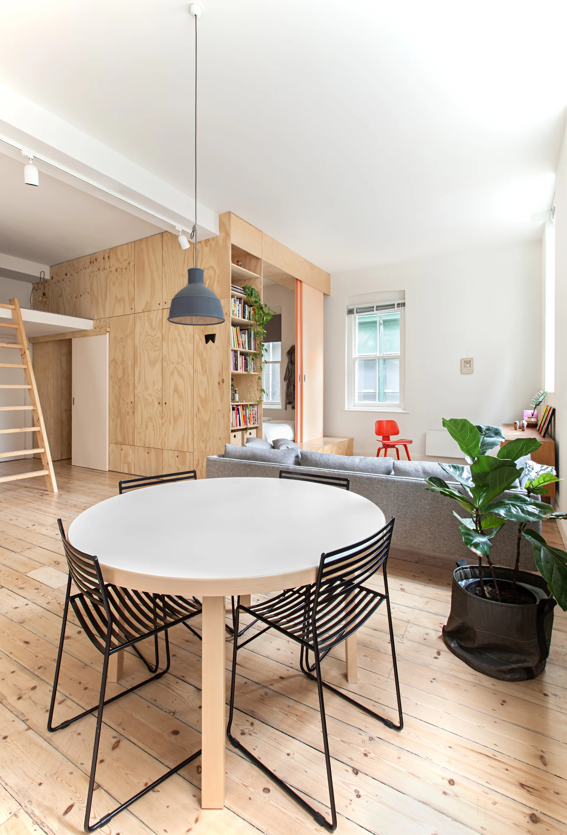 An open-plan living and dining space features a round white table, a grey sofa, and a large plywood storage wall with a loft ladder.