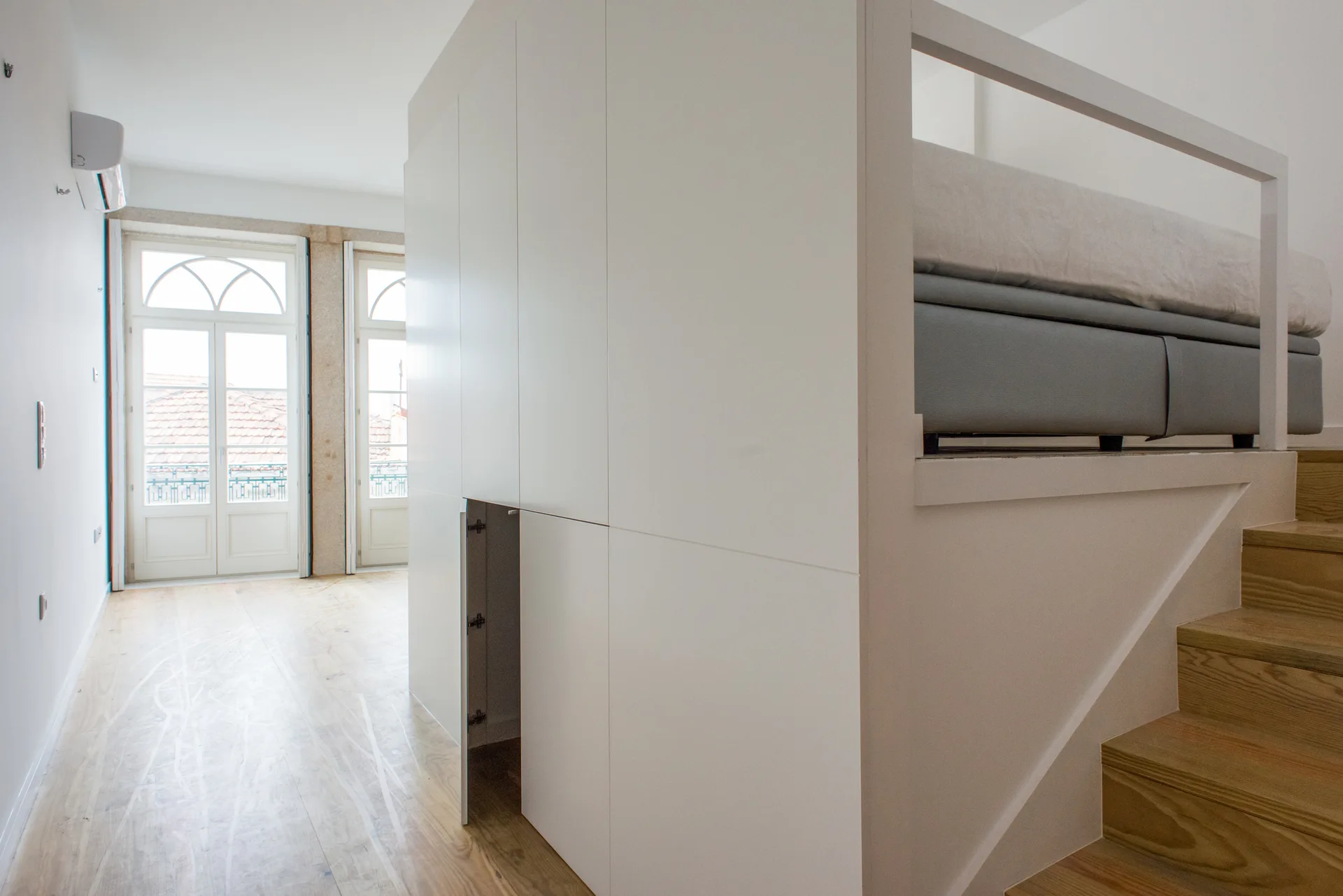 A modern loft bed rests atop a white built-in storage unit with wooden stairs leading up, set in a minimalist room with hardwood floors.