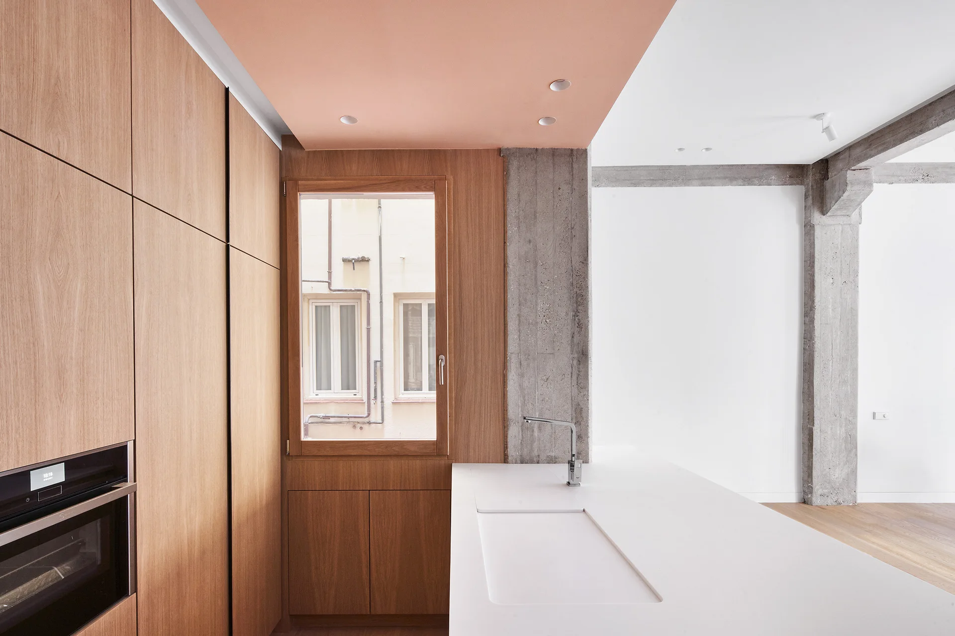 A minimalist kitchen features flat-panel wood cabinets, a white island with an integrated sink, an exposed concrete column, and a peach ceiling.