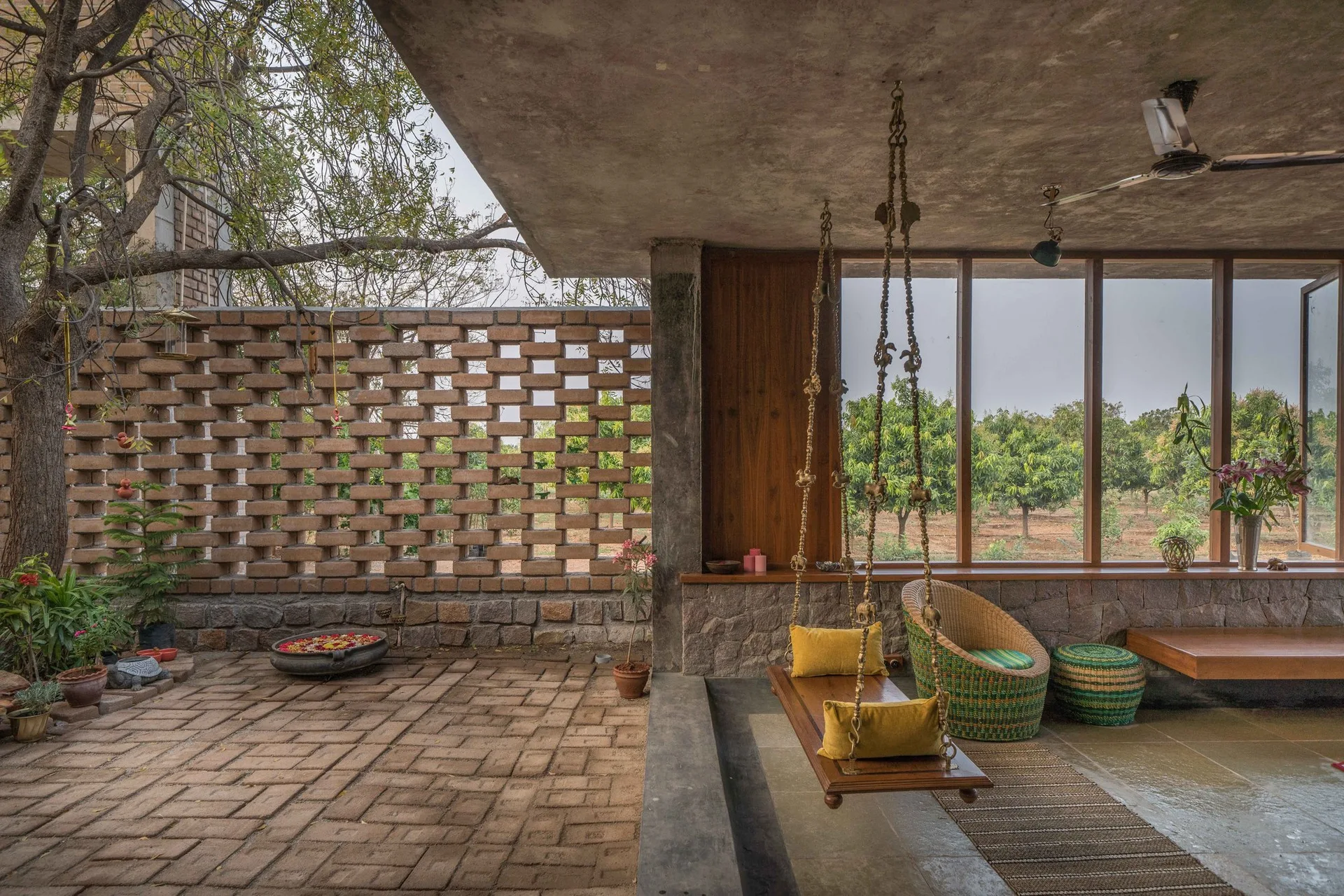 A covered outdoor patio featuring a hanging wooden swing with yellow pillows, a brick lattice screen, a wicker chair, and large windows looking onto a garden.