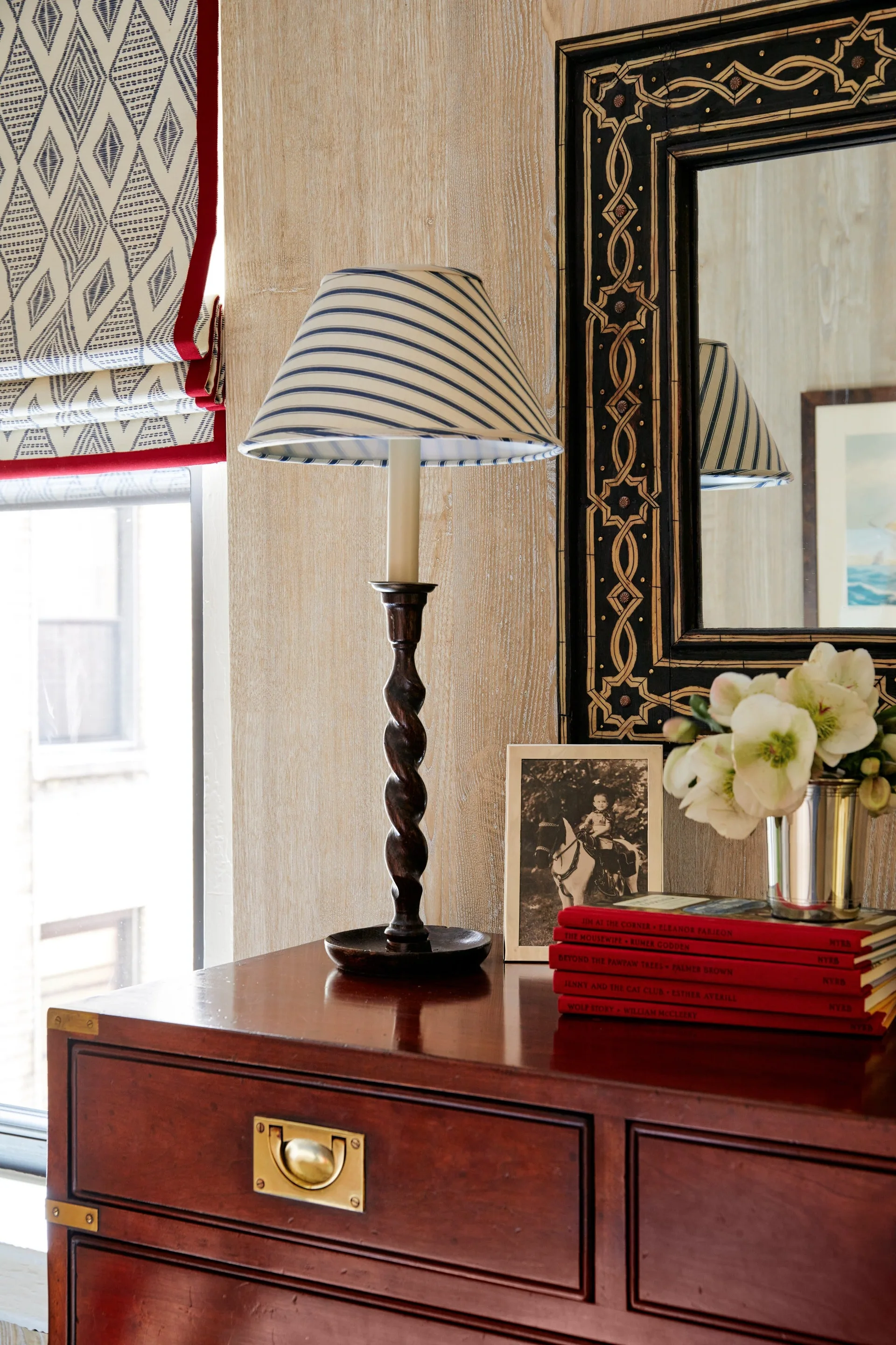 A dark wood campaign chest features a twisted wooden lamp with a striped shade beneath a black patterned mirror on light wood paneled walls.