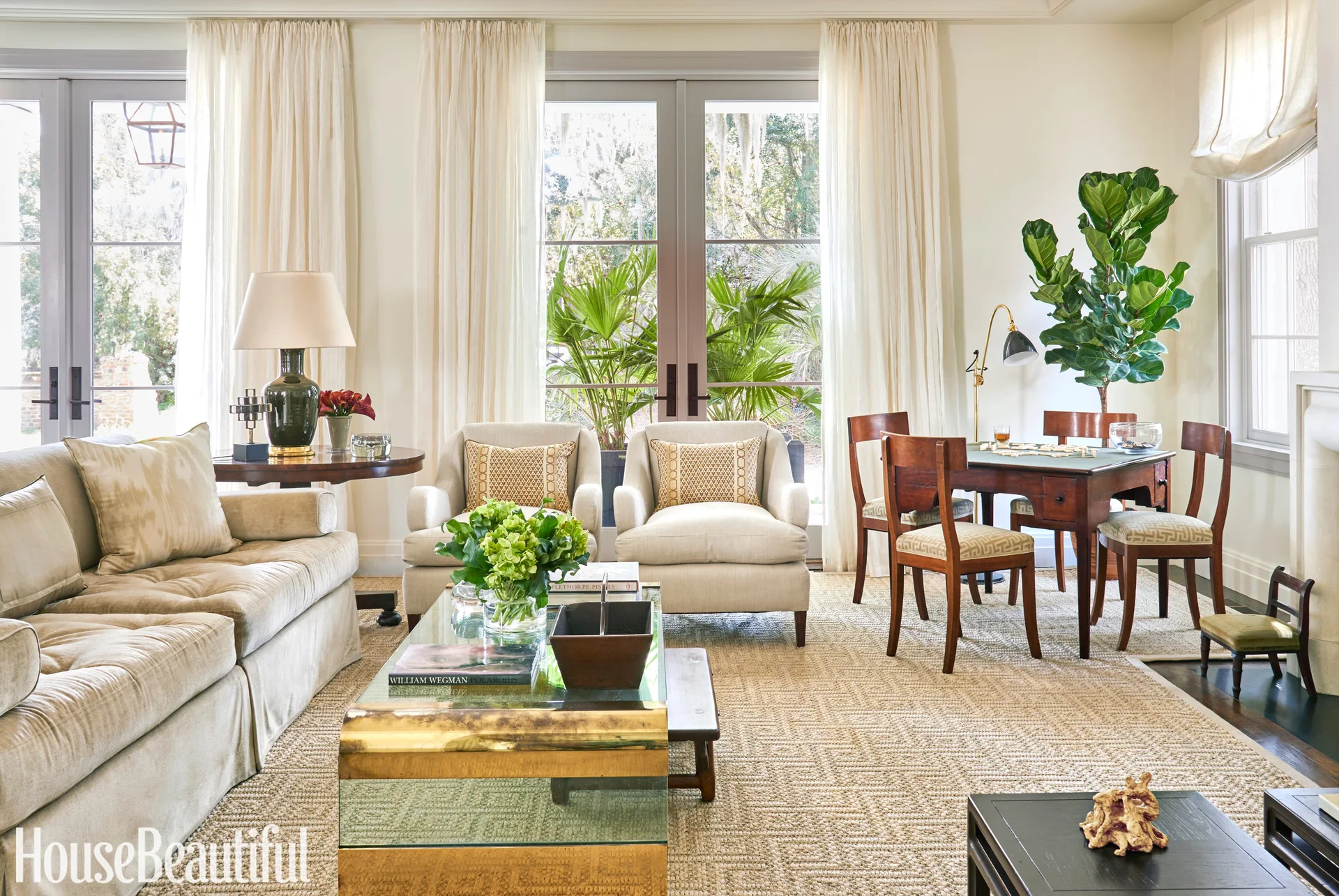 A sunlit cream living room with two upholstered club chairs, a brass and glass coffee table, a wooden game table, and sheer white curtains.
