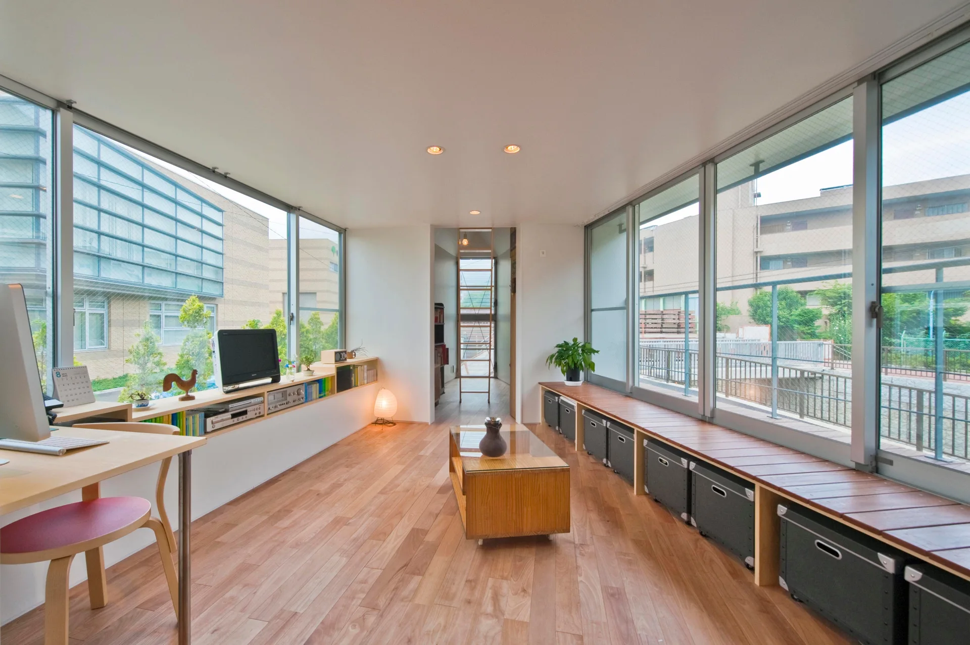 A minimalist wood room features dual window walls, a central wood coffee table, and a long built-in bench lined with grey fabric storage bins.