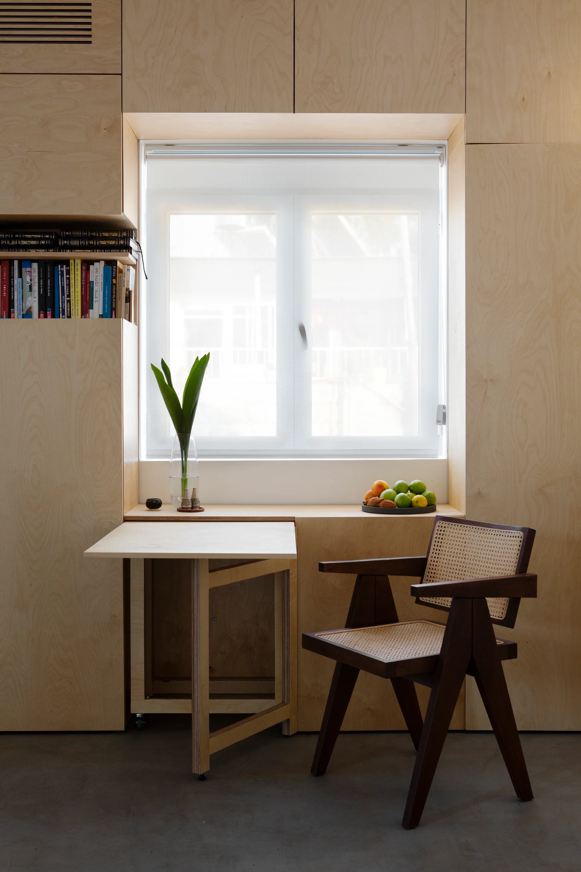 A minimalist light wood built-in window nook features a drop-leaf folding desk and a dark wood chair with a rattan seat and back.