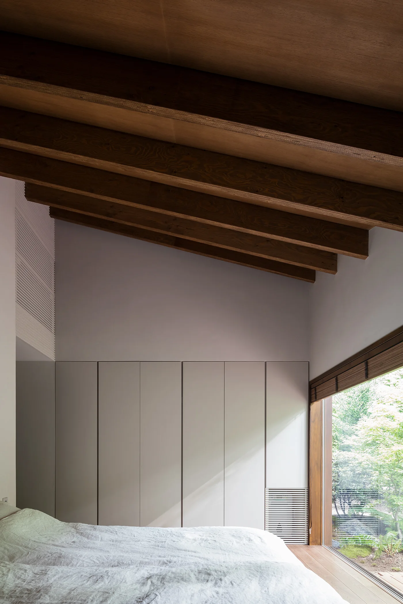 A minimalist bedroom featuring light grey built-in wardrobes, a sloped ceiling with exposed dark wood beams, and a large window looking out to greenery.