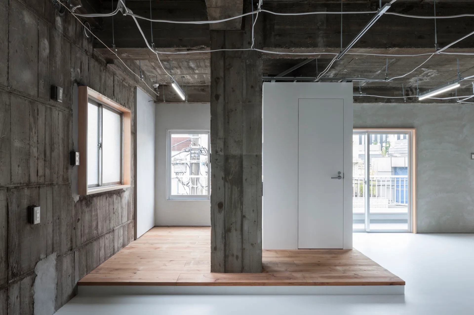 A minimalist room features a central concrete column, an exposed concrete ceiling with wiring, and a raised wooden platform with a white partition door.
