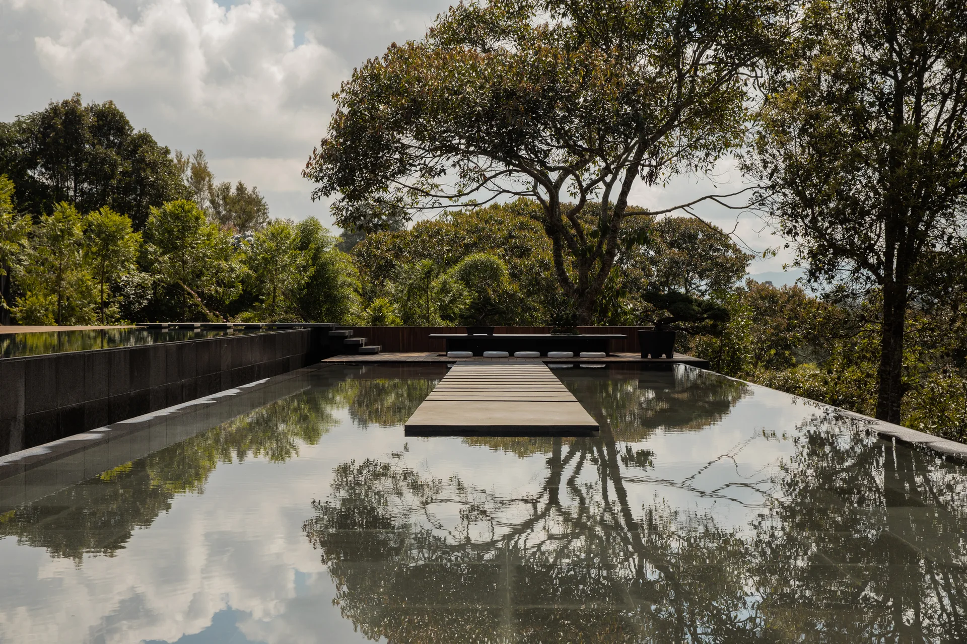 A large reflective pool features a raised concrete stepping stone pathway leading to a wooden deck with a low bench under a mature tree.