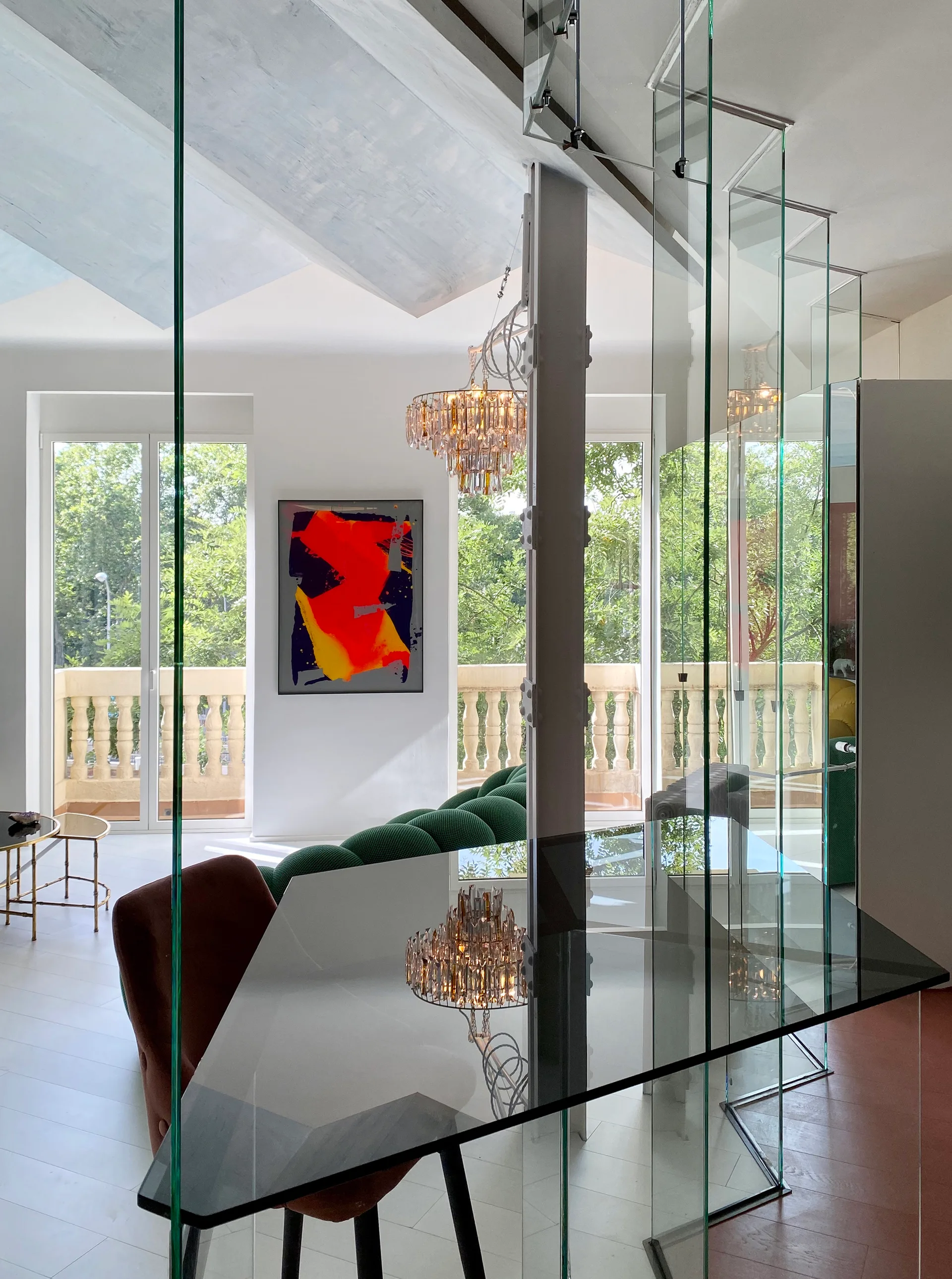 A modern dining area features a dark glass table next to a transparent glass partition, looking into a living room with a green tufted sofa.