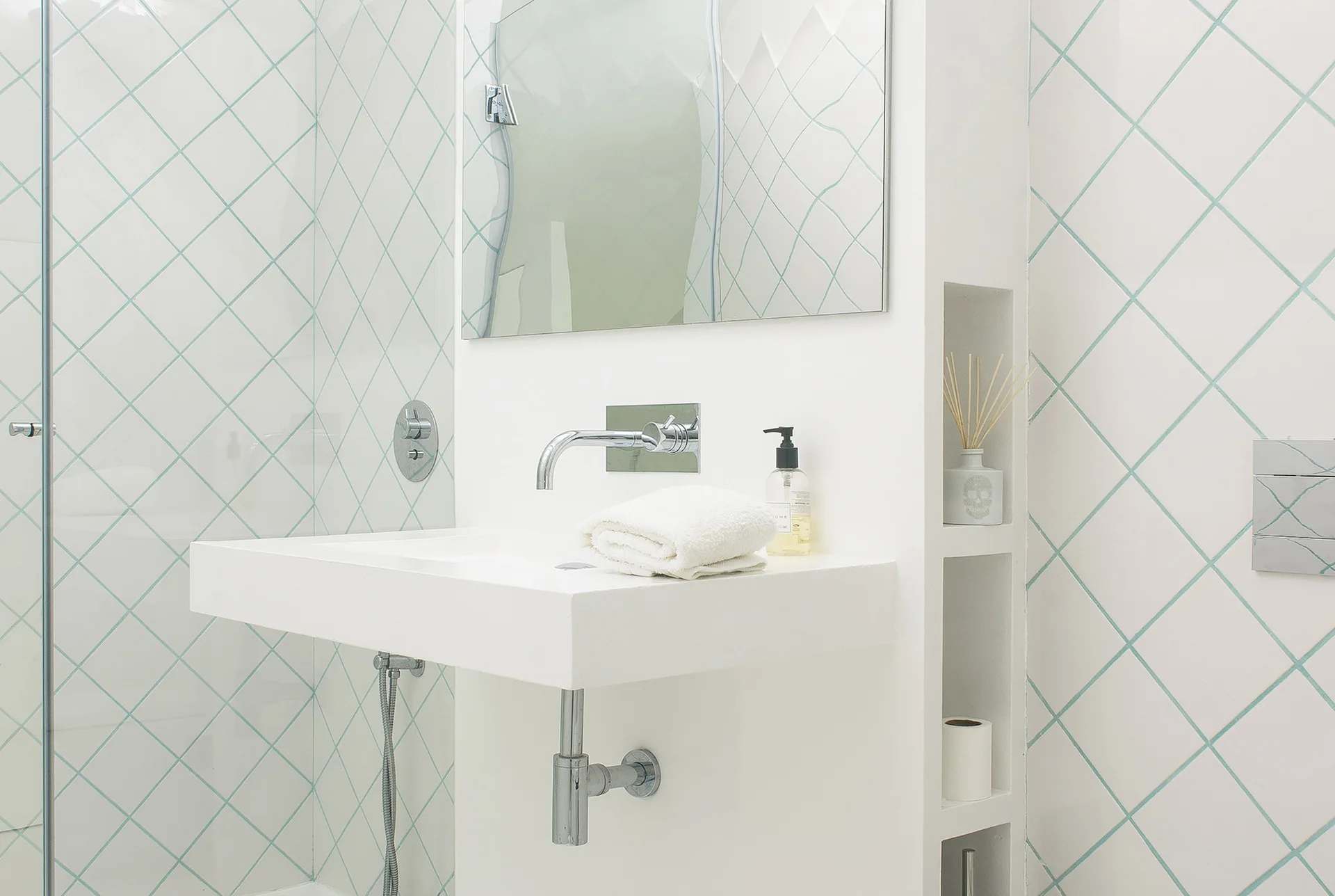 A modern white wall-mounted sink features a chrome faucet set against a white tiled wall with diamond-shaped turquoise grout lines.