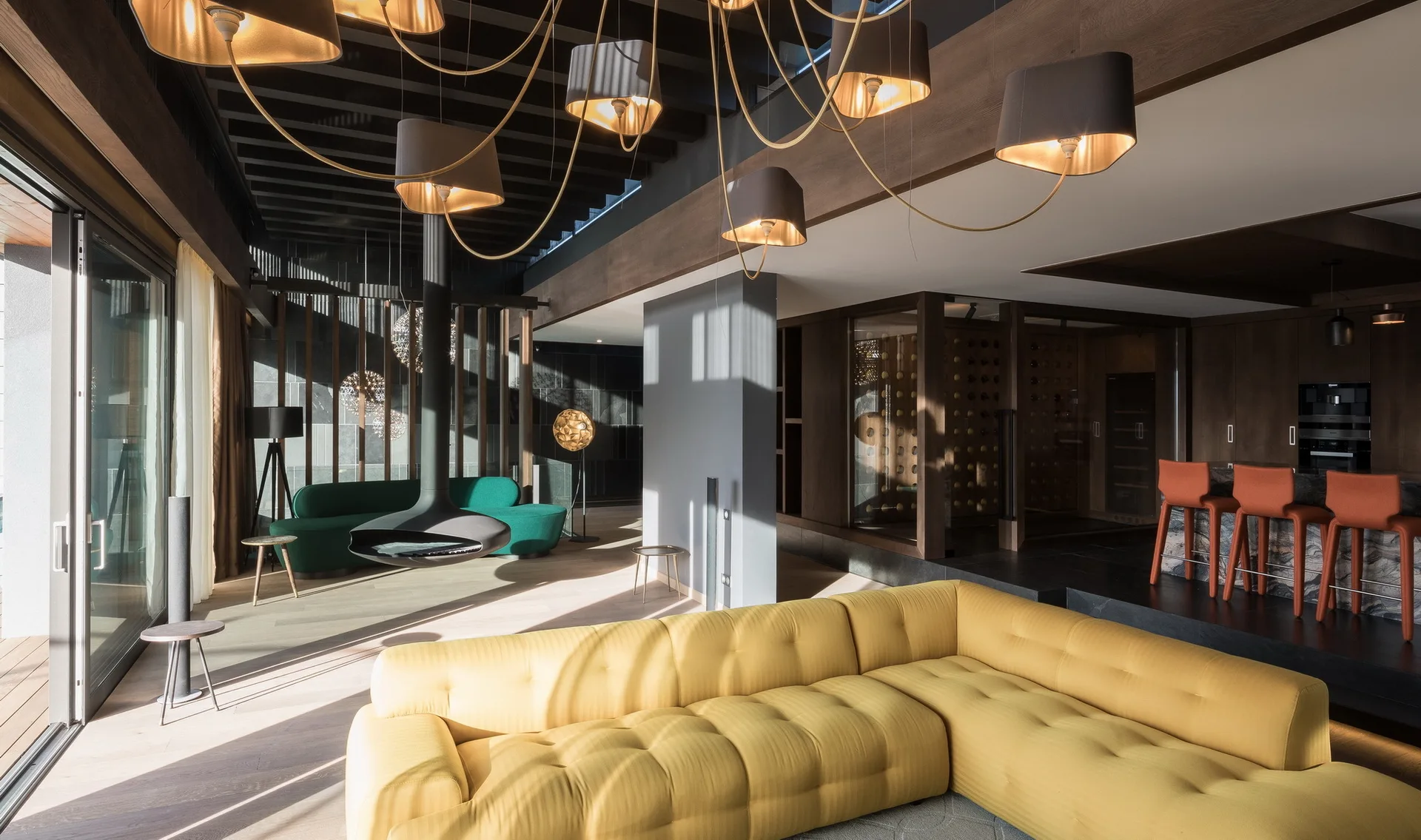 A contemporary open-plan living room features a tufted yellow sectional sofa, a black suspended fireplace, and a glass-enclosed wine cellar.
